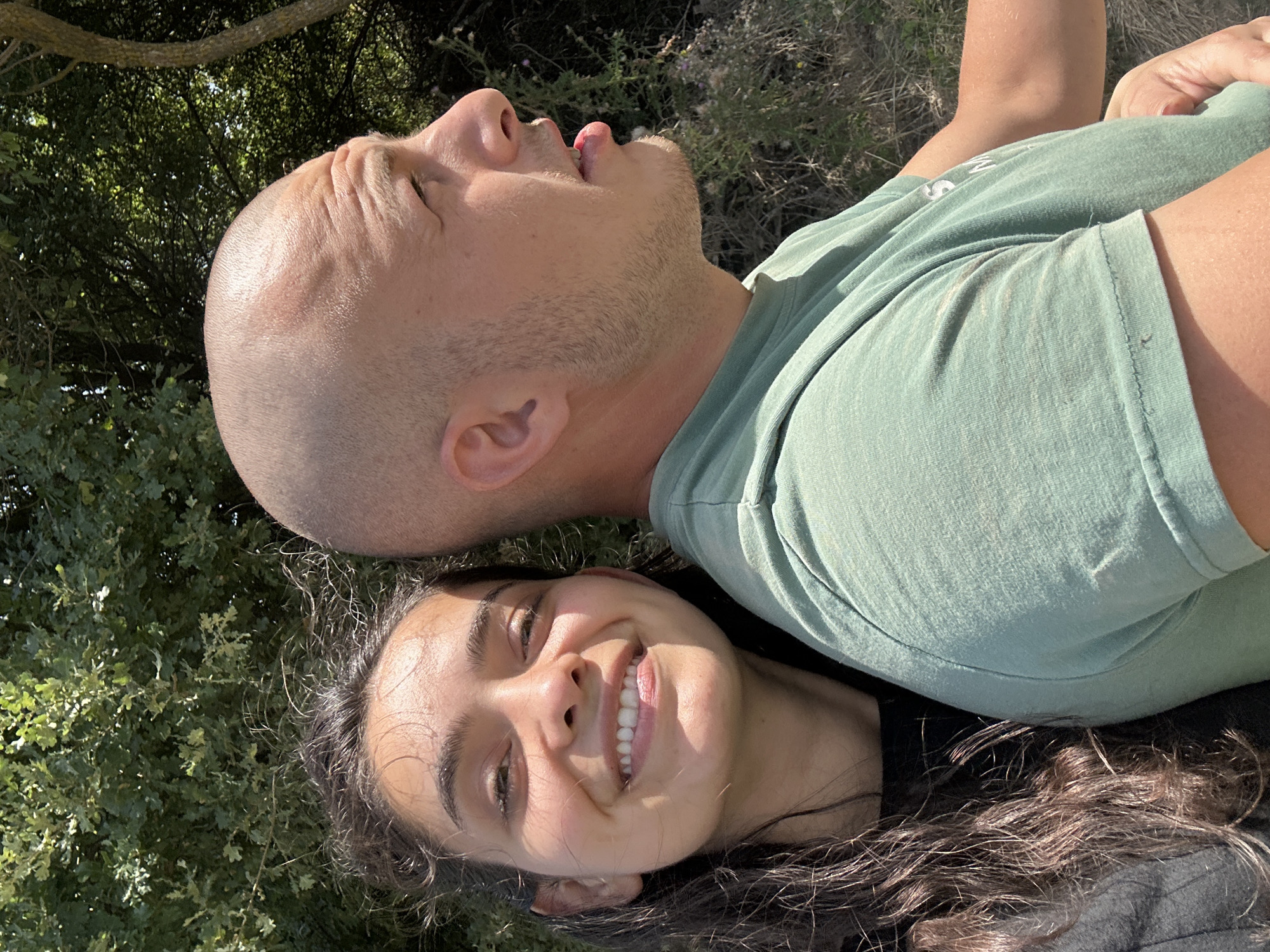 The image shows a close-up, side-by-side portrait of a young woman and a man. The woman is on the left, facing the camera and smiling. She has long, dark, wavy hair and appears to be wearing a dark shirt. The man is on the right, with a shaved head and is looking to his right. He is wearing a light green t-shirt. They are positioned outdoors with a backdrop of lush green trees and foliage, suggesting a natural environment. The lighting indicates it is daytime, likely sunny. The image appears to be taken in Malevo, Bulgaria, though no specific landmarks are visible to confirm this.