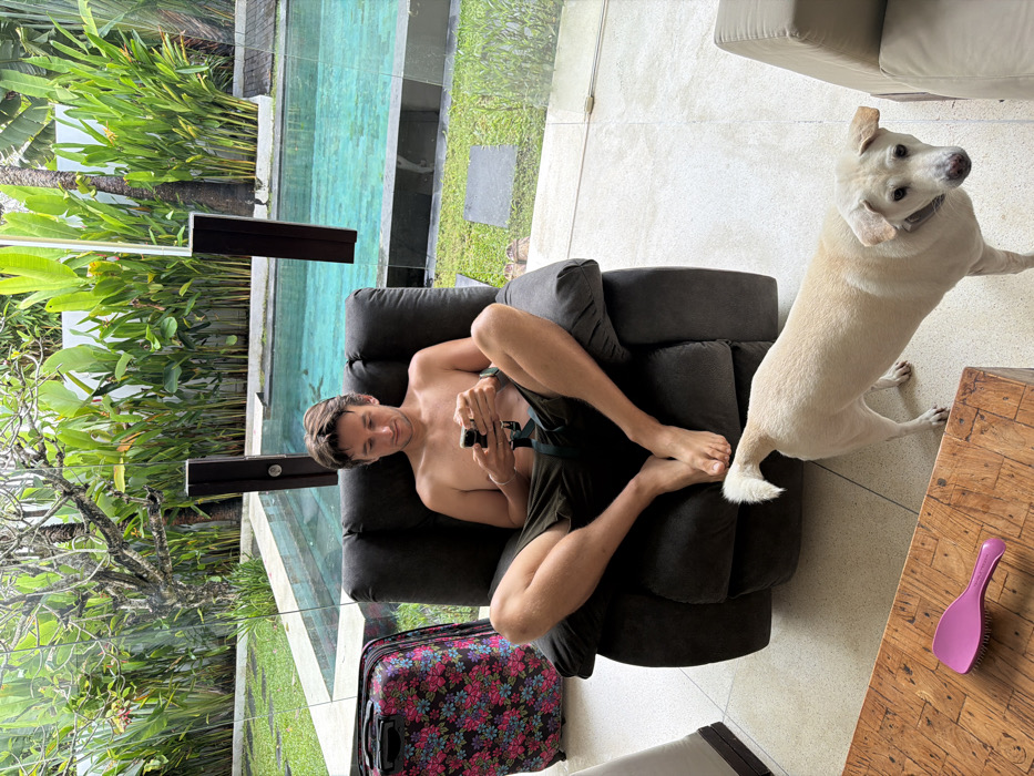 The scene depicts an interior space overlooking an outdoor area at a location in Ungasan, Indonesia. An adult male is seated in a dark grey recliner, bare-chested and wearing dark shorts, with his bare feet resting on the light-colored tiled floor. He is actively using a dark-colored smartphone held in both hands, looking downwards at the device. A floral-patterned suitcase, predominantly pink, blue, and green on a dark background, stands upright to the left of the recliner. A light-colored, short-haired dog with a light collar stands on the tiled floor in the foreground, positioned to the right of the male's feet and looking towards the viewer. A wooden coffee table with a textured top is partially visible in the lower left, on which a bright pink detangling hairbrush is placed. A large, clear glass wall, incorporating dark vertical structural elements, separates the indoor space from an outdoor area. Beyond this glass, a swimming pool with light blue water is visible, surrounded by green grass, dark paving, and dense tropical vegetation, including large-leafed plants and trees.