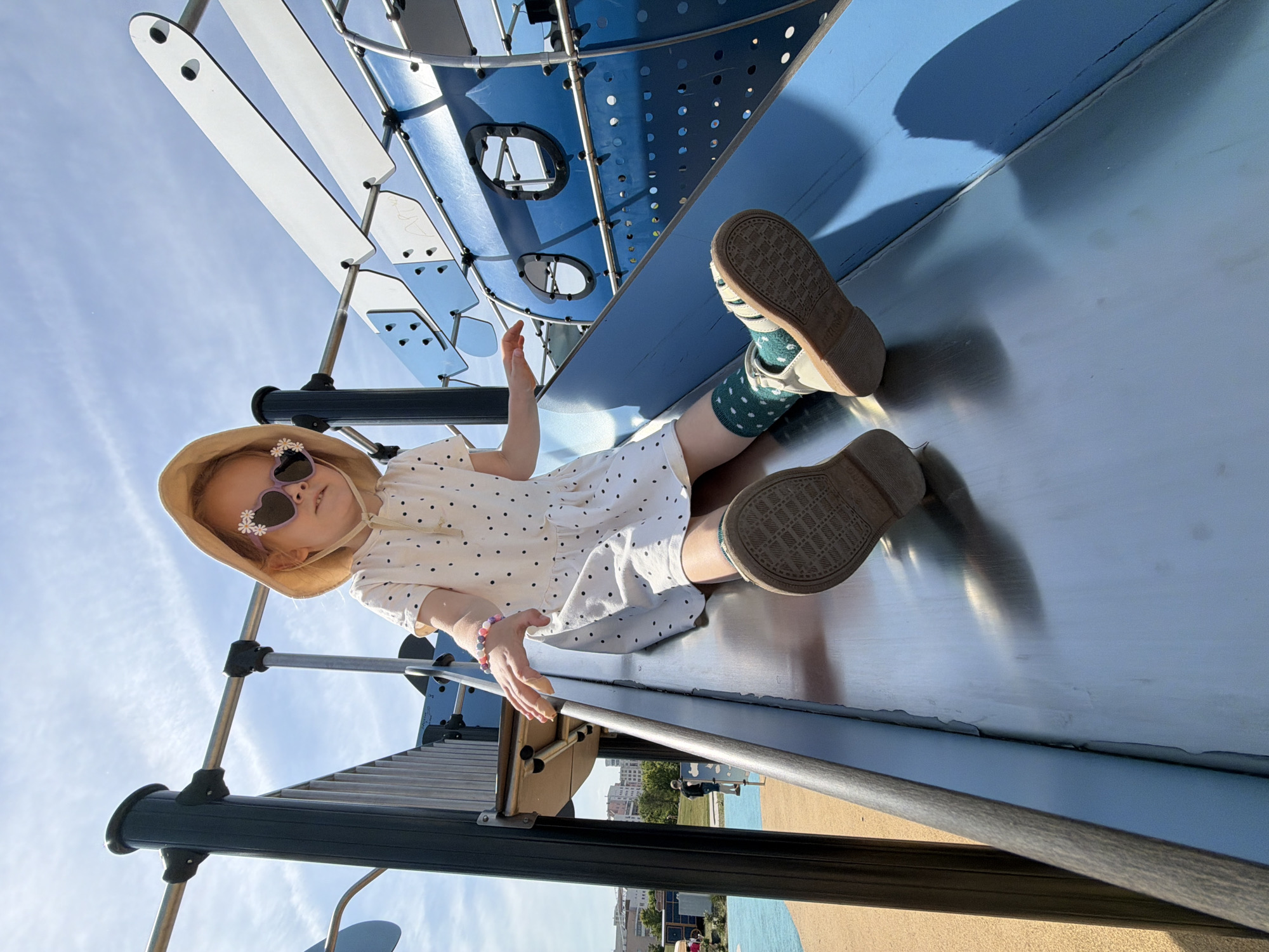 A young child is depicted in a low-angle, eye-level shot, actively descending a metal playground slide. The child wears a beige wide-brimmed bucket hat and light purple heart-shaped sunglasses detailed with small white daisies on the frames. Her attire includes a white short-sleeved dress patterned with small black polka dots, green socks with white polka dots, and light-colored sandals with textured brown soles. A multi-colored beaded bracelet is visible on her left wrist. Her hands are slightly raised, and her gaze is directed forward. The slide is constructed from a silvery metal, reflecting sunlight, and is integrated into a larger playground structure featuring dark gray vertical support beams and a prominent blue, perforated panel with two circular window-like cutouts. Additional white and blue panels are visible at the top of the structure. The ground immediately adjacent to the slide consists of a tan, possibly rubberized or sand-filled, surface. In the background on the left, an indistinct human figure is partially visible. Further back, buildings of an urban environment and green foliage are present beneath a clear blue sky with sparse white cloud formations, indicating fair weather. The scene is illuminated by strong direct sunlight, casting distinct shadows. The location is the city of Khimki, Russia.