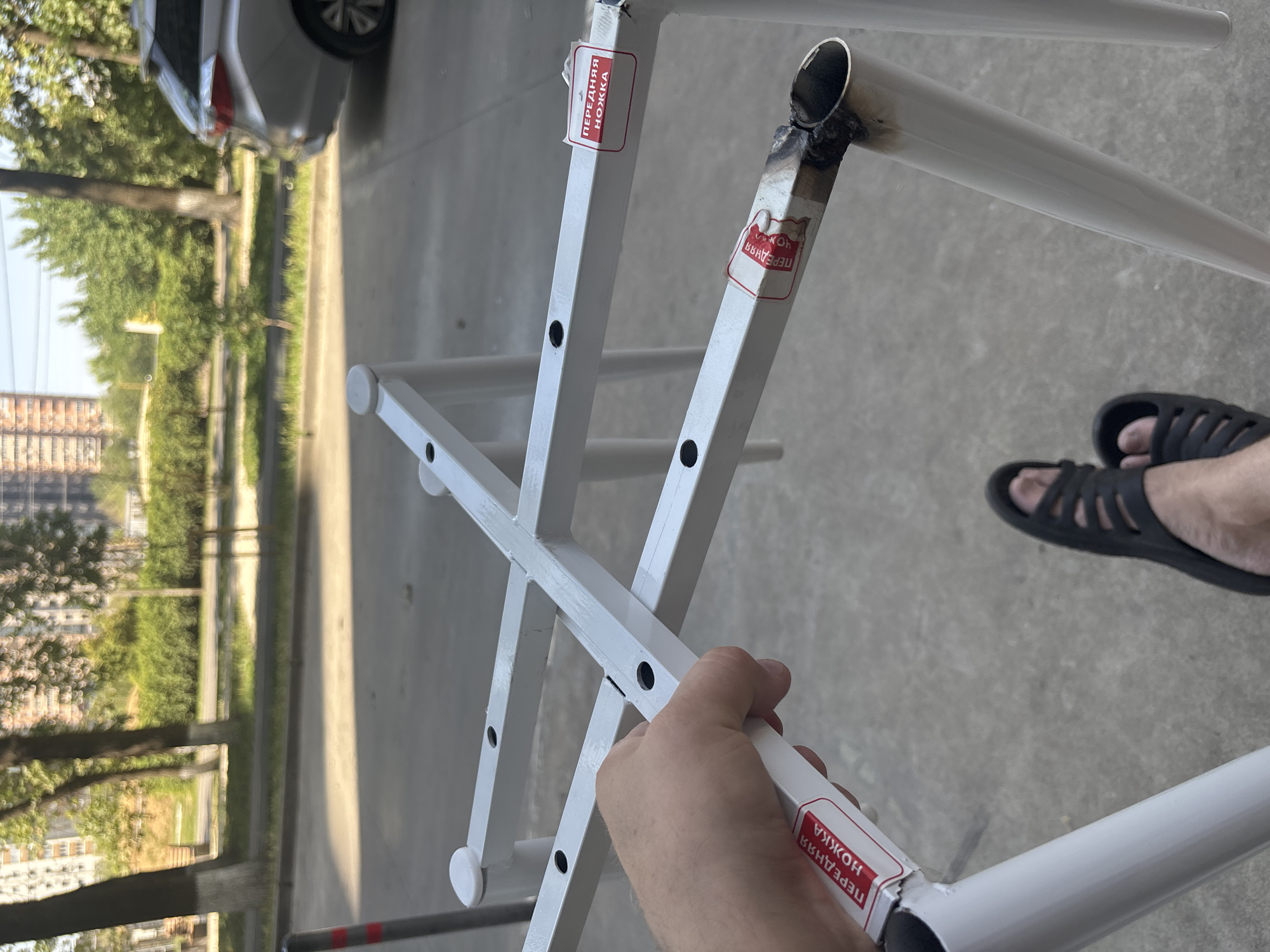 A first-person perspective shot shows a person's left hand holding a white metal frame, possibly a part of a folding table or chair, which has a sticker on it reading "ПЕРЕДНЯЯ НОЖКА" (front leg) in red lettering. The frame is white, with some visible weld marks and a blackened, melted section on one of the pipes. In the background, a paved surface transitions to a grassy area with trees and buildings. A silver car is parked on the right side of the image. A person's feet wearing black sandals are visible at the bottom of the frame. The image was taken outdoors during the day, with sunlight casting shadows. The scene suggests an urban environment, possibly a residential area, though no specific landmarks in Krasnodar, Russia, are identifiable.