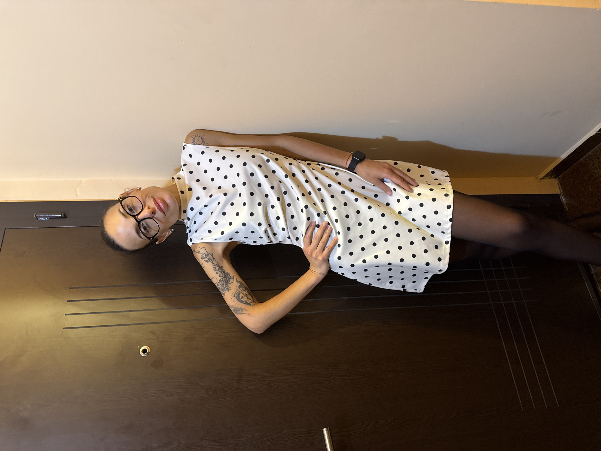 A woman with a shaved head and glasses is lying on a dark wooden door in an interior setting. She is wearing a white dress with black polka dots and black tights. Her arms are positioned on the dress, and there are tattoos visible on her arms. The door is in a building in the city of Pleven, Bulgaria. The background consists of a light beige or off-white wall and a glimpse of another dark wood door in the upper right corner.