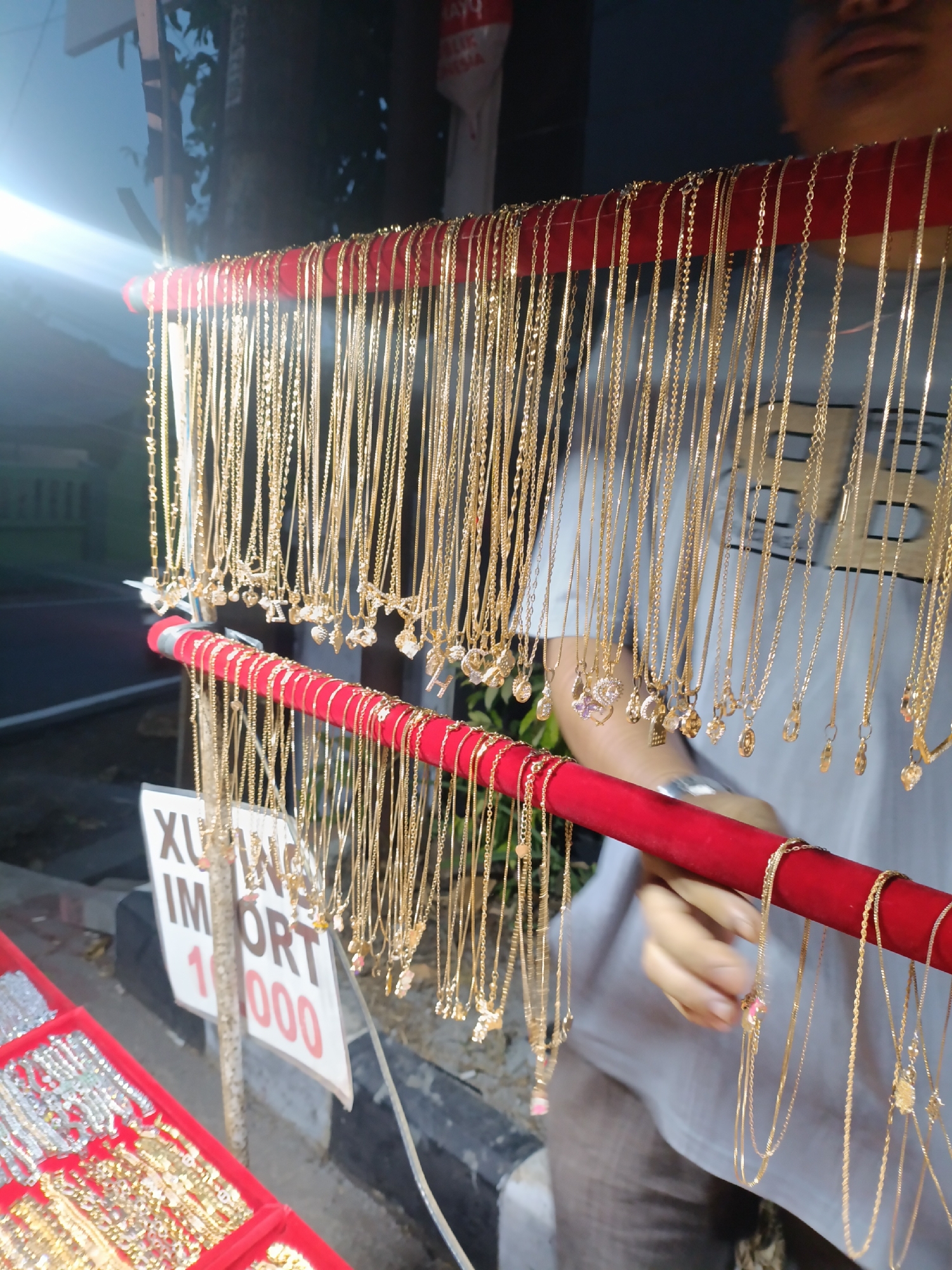 In Jonggol, Indonesia, a street vendor displays an assortment of gold necklaces on a red display rack. The necklaces, varying in style and chain thickness, are hung in multiple rows. Some necklaces feature small pendants, including letters and what appear to be small gemstones. A white sign with red lettering reading "IMPORT 1000" is visible below the necklaces. In the foreground, a red tray holds other jewelry items, possibly bracelets and necklaces. A person, partially visible, is interacting with the displayed necklaces, with their hands reaching towards them. The background suggests an outdoor setting with trees and a road, indicating a typical street-side market environment.
