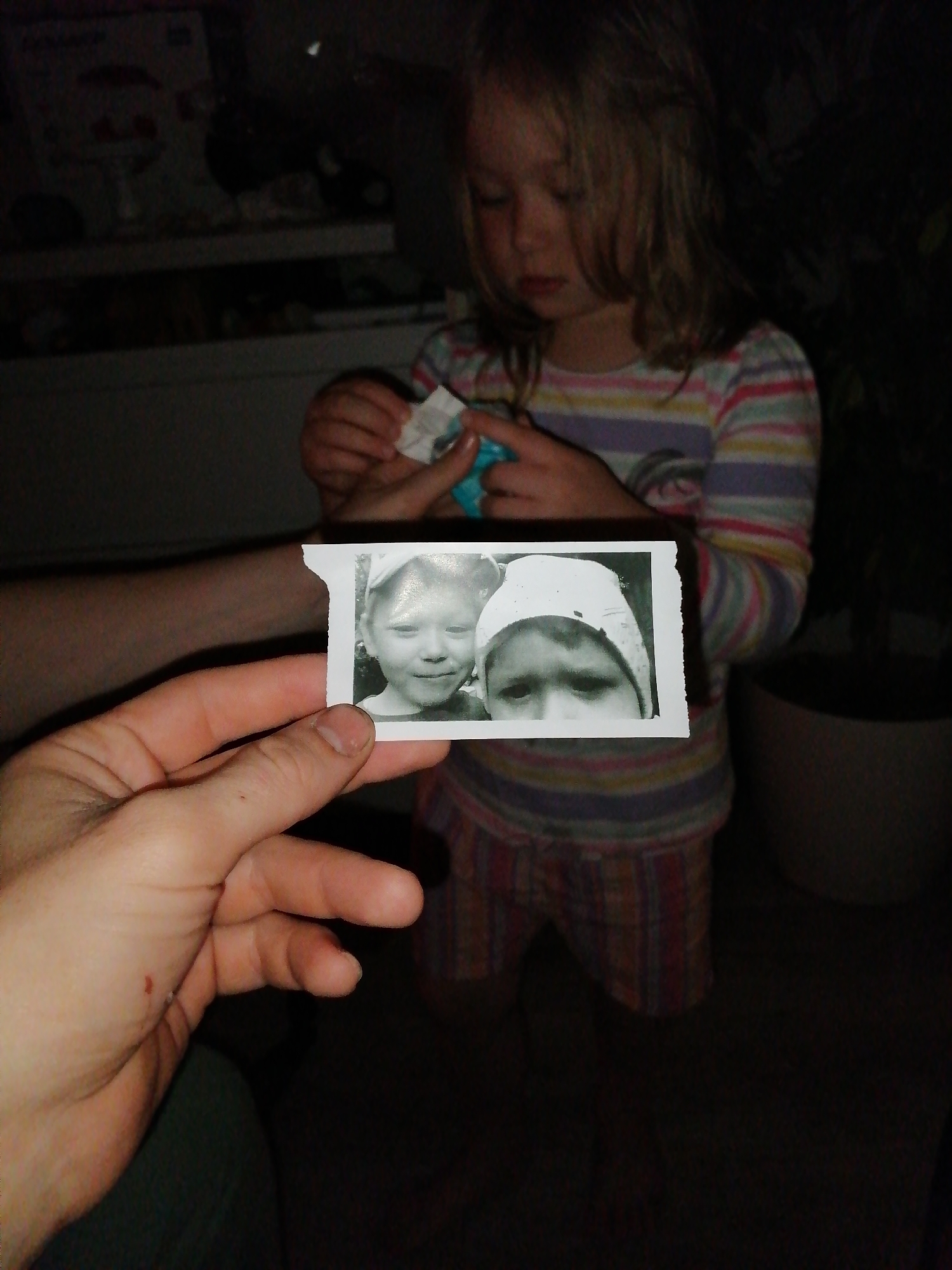 A young child with blonde hair, wearing a striped long-sleeved shirt and plaid shorts, stands in a dimly lit room. The child is holding a small, black and white photograph in their hands. The photograph shows two children's faces, one on the left and one on the right. A person's hand is visible in the foreground, holding the photograph. In the background, shelves with various items are partially visible, suggesting an indoor environment. The lighting is low, creating shadows and a dark atmosphere. The location is Unknown, Unknown.