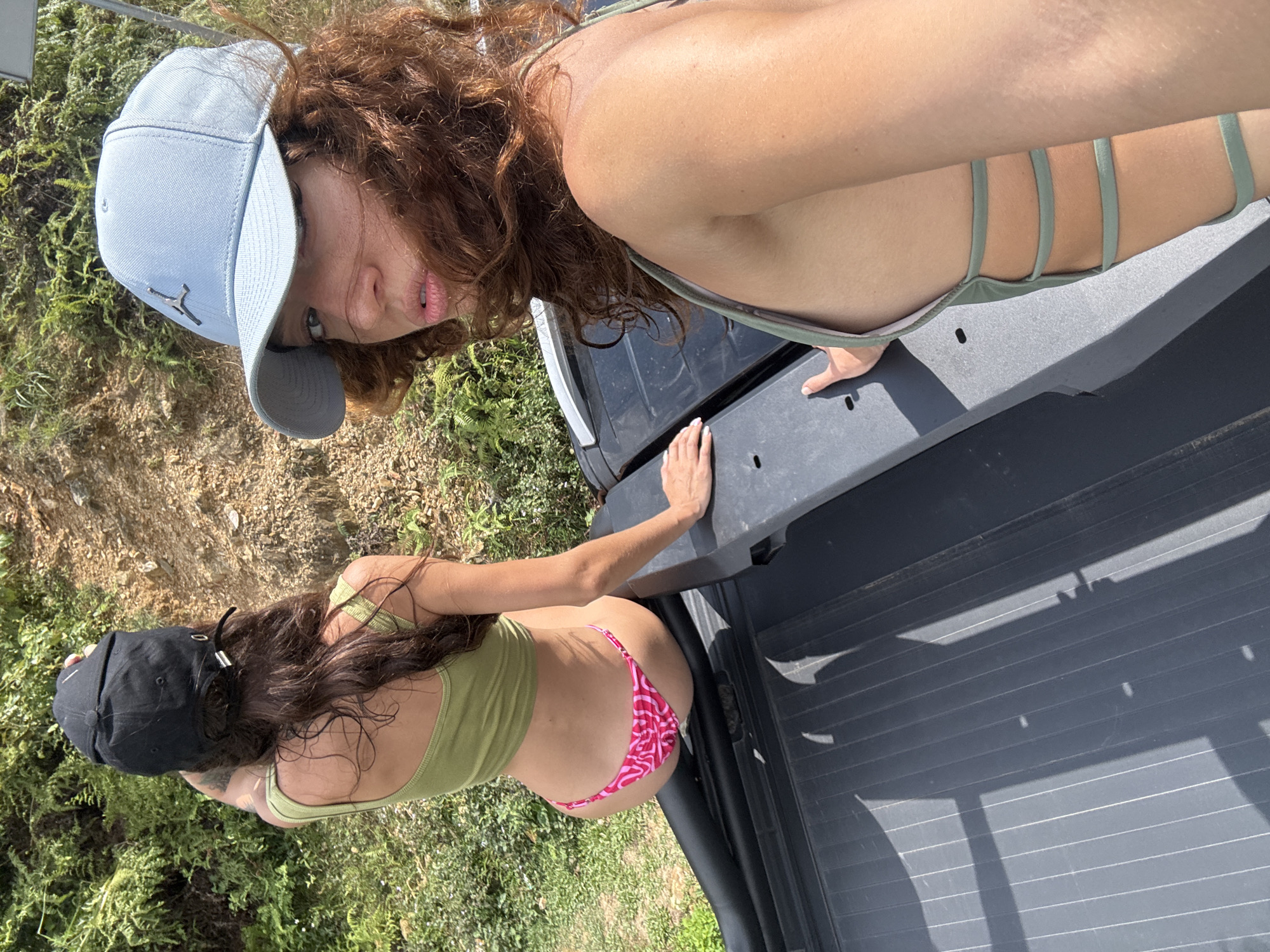 Two women are posing on the tailgate of a pickup truck. The woman at the top of the frame is wearing a light blue baseball cap and a light green swimsuit top with straps. She has curly red hair. The other woman is wearing a black baseball cap, a light green swimsuit top, and pink and white bikini bottoms. The tailgate of the truck is black and ribbed. The surrounding environment is grassy with some small trees. The location is unspecified.