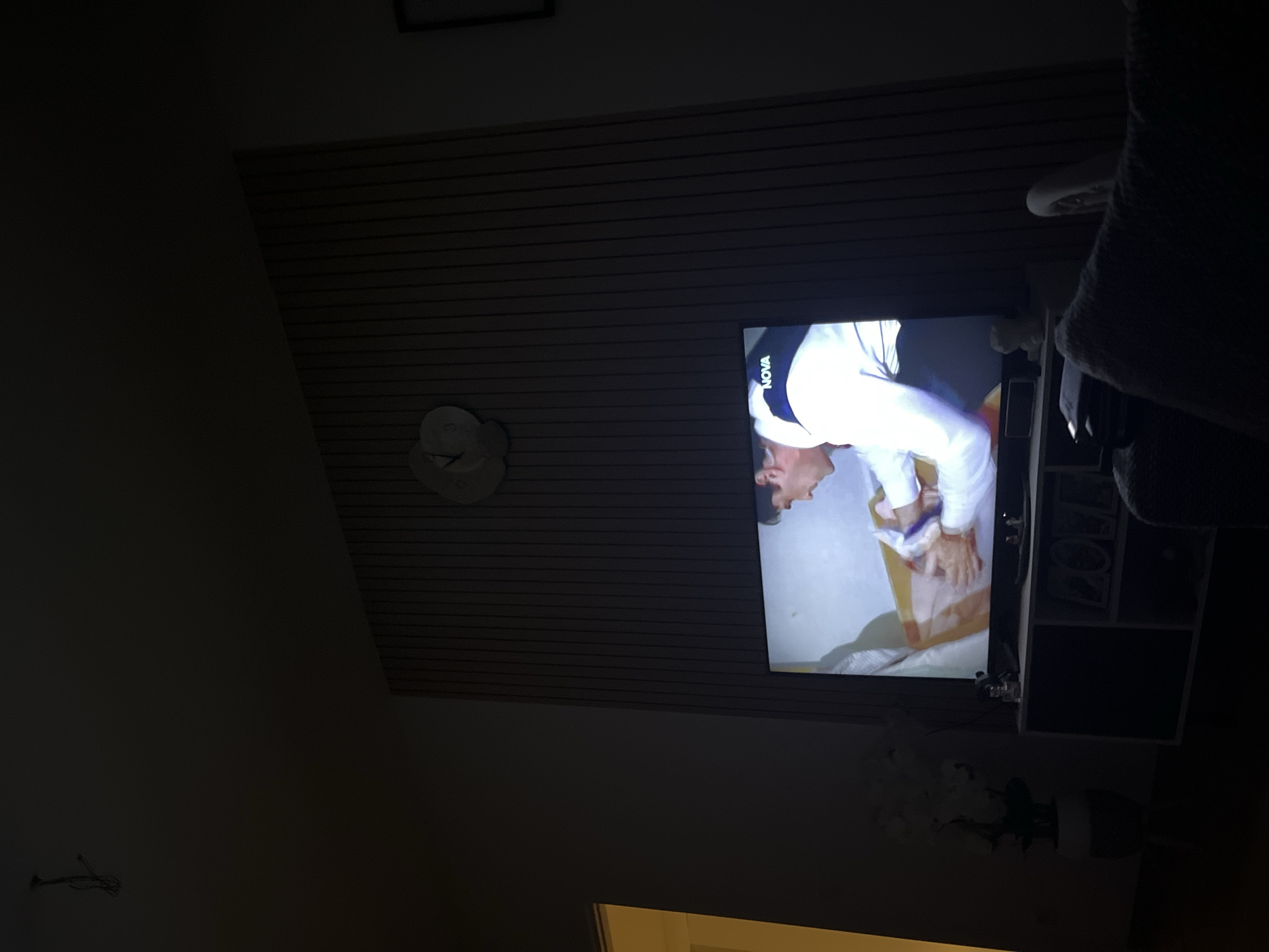 The image shows a dimly lit living room with a television screen displaying a scene. On the screen, a man in a white shirt and blue collar is seen handling a cardboard box. The television is placed on a dark entertainment stand. To the left of the TV, a potted plant with white flowers is visible. The background features a wall with vertical wooden slats and a decorative clock. The overall impression is of a modern living space. The location is the city of Garmisch-Partenkirchen, Germany.