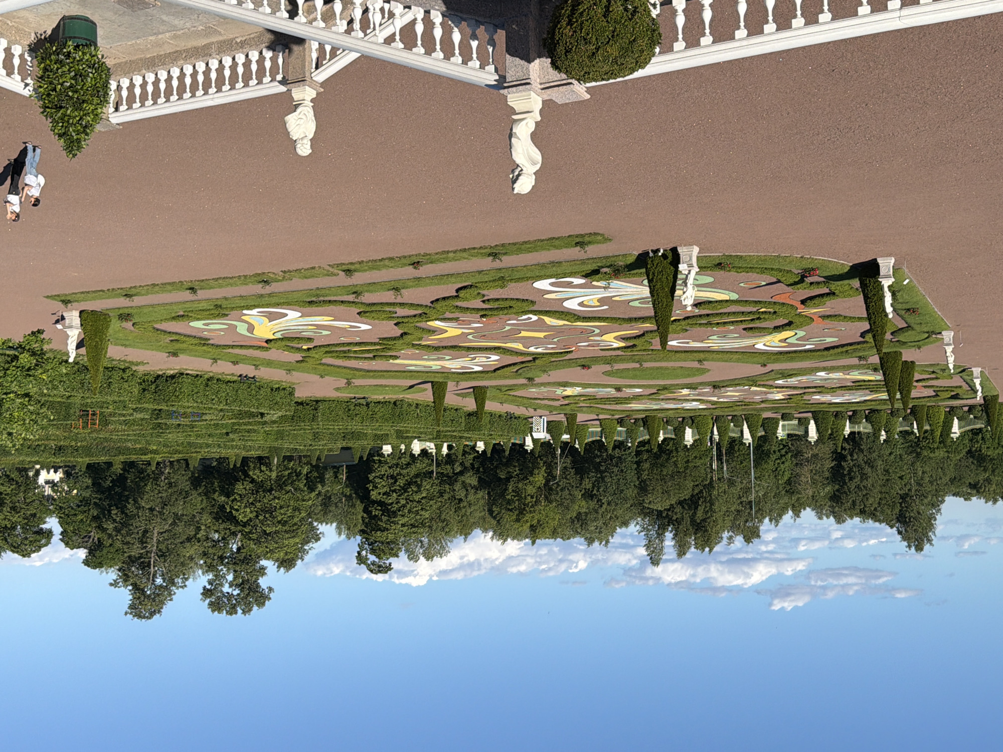 An exterior, eye-level, full shot captures a formal garden under a bright, blue sky with scattered white clouds. The garden features a large, geometrically patterned flowerbed in the center, with swirling designs of green hedges, white gravel, and colorful flower arrangements in red, yellow, and blue. Flanking the flowerbed are evenly spaced, dark green, conical trees. In the foreground, a white balustrade lines a path, and on top of it are two busts. To the right of the busts, a potted, dark green, conical tree sits in a green planter. Two individuals, a woman and a man, are walking on a brown gravel path in the middle right of the image. Behind the flowerbed is a dense line of manicured green hedges, forming rectangular sections. Further in the background, a line of trees forms a backdrop, with some scattered white statues visible. The scene is well-lit by natural daylight.