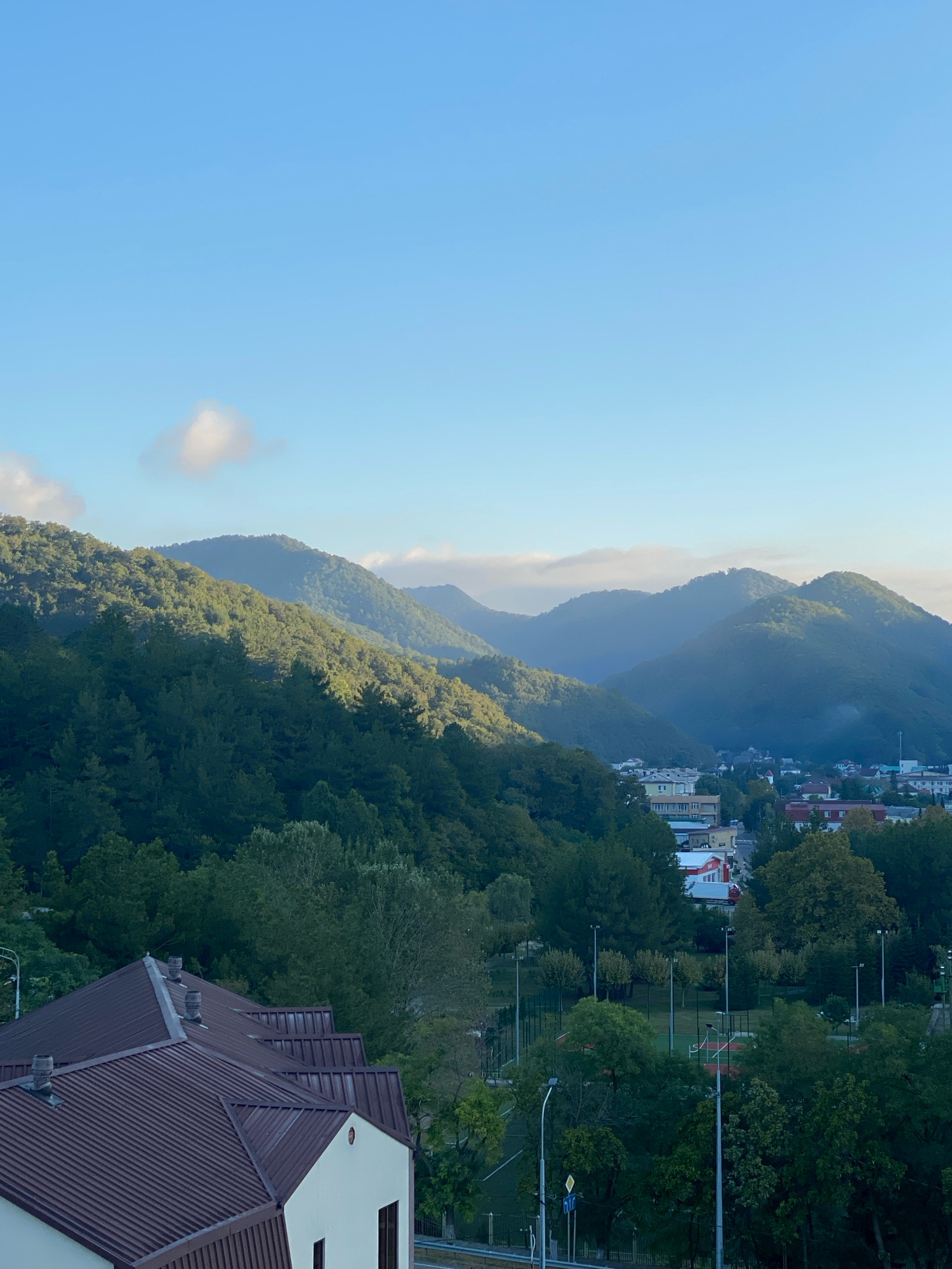 The image depicts a panoramic view of the city of Nebug, Russia, nestled amidst lush green mountains. The foreground is dominated by the brown, corrugated metal roofs of several buildings, one of which has a beige facade and a visible window. Beyond the buildings, a sports complex with a tennis court and visible lampposts is nestled among trees. The mid-ground shows a town with various buildings, including some with red roofs, scattered among the dense green foliage. The background is characterized by rolling, tree-covered mountains that recede into the distance, with the sky above transitioning from a clear blue at the top to a hazy lighter blue near the horizon. A few wispy clouds are visible in the sky. The lighting suggests it is either early morning or late afternoon, with sunlight casting shadows and highlighting certain slopes of the mountains. There are no people or animals visible in the image.