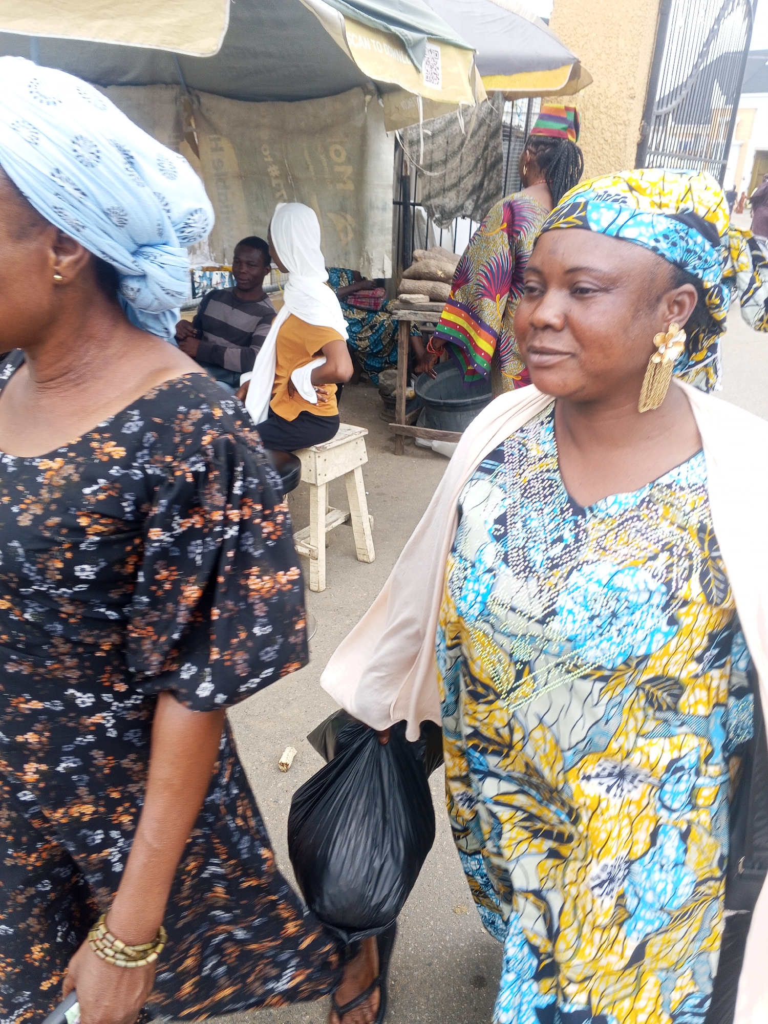 A medium shot captures a street market scene, with three women in the foreground. The woman on the right, in focus, wears a brightly patterned blue, yellow, and black dress with a light pink overcoat and large gold tassel earrings. She carries a black plastic bag in her left hand. To her left and slightly behind, another woman in a dark floral dress is partially visible, also holding a black bag. A third woman, in a white headscarf and an orange top, sits on a stool in the background. A man in a striped shirt is seated next to her. The environment is a bustling marketplace, with stalls and awnings visible, suggesting a public outdoor setting. The ground is paved and appears to be asphalt.