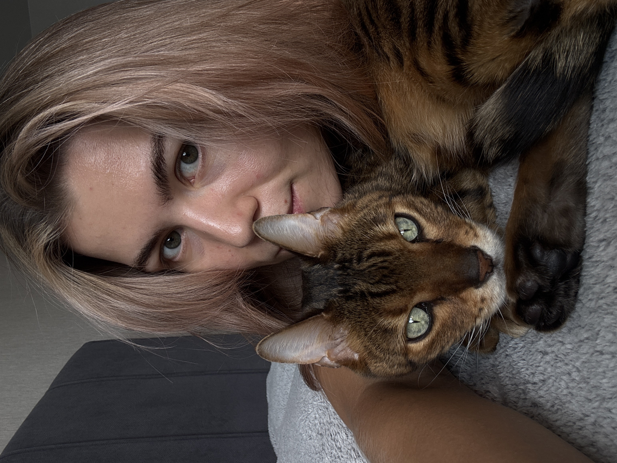 A close-up, rotated shot features a person and a cat. The person, with light blonde-brown hair and fair skin, has light green eyes and a small smile. The cat, a tabby breed, has green eyes and brown-black fur with striped markings. The cat is lying on the person. The background includes a gray blanket and a dark gray surface. The location is unknown.