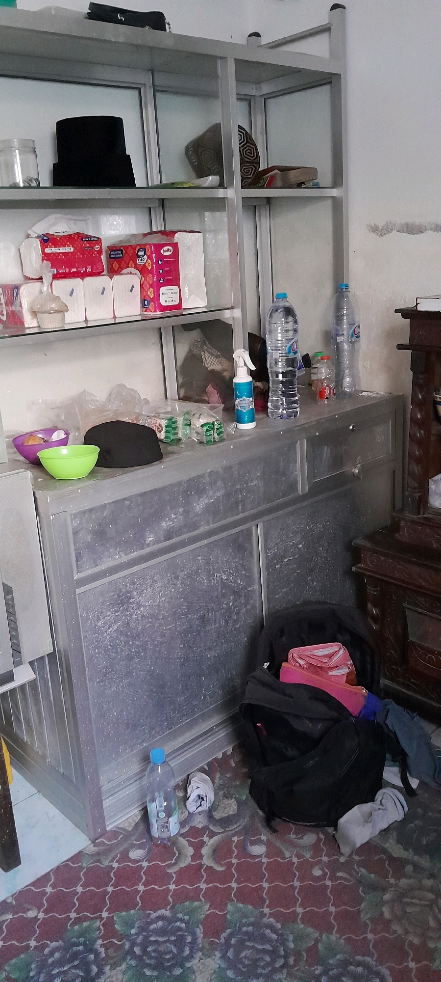 The image depicts an indoor scene, likely a storage area or living space, with an emphasis on shelves and a metal cabinet. The shelves, constructed of metal and glass, are partially filled with various items. On the top shelf, a black hat or cap rests next to a clear glass jar. Below this, several packets of tissues and other packaged goods are organized. The middle shelf contains a purple bowl, a green bowl, a black item that appears to be a hairpiece or wig, and several small packages. A spray bottle and two large plastic water bottles are positioned on the top of the metal cabinet. The cabinet itself is metallic grey with visible dust and smudges. On the floor, a black backpack is open and contains folded pink and blue fabric. Several white cloth items are scattered around the backpack and a single plastic water bottle stands upright on the floor. The flooring is covered by a patterned rug with floral designs in shades of red and dark purple. The background wall is painted white, with some visible marks and discoloration. The lighting suggests an indoor environment, possibly with ambient light from a window or artificial sources. No explicit geographical markers for Malang, Indonesia are visible within the image itself.