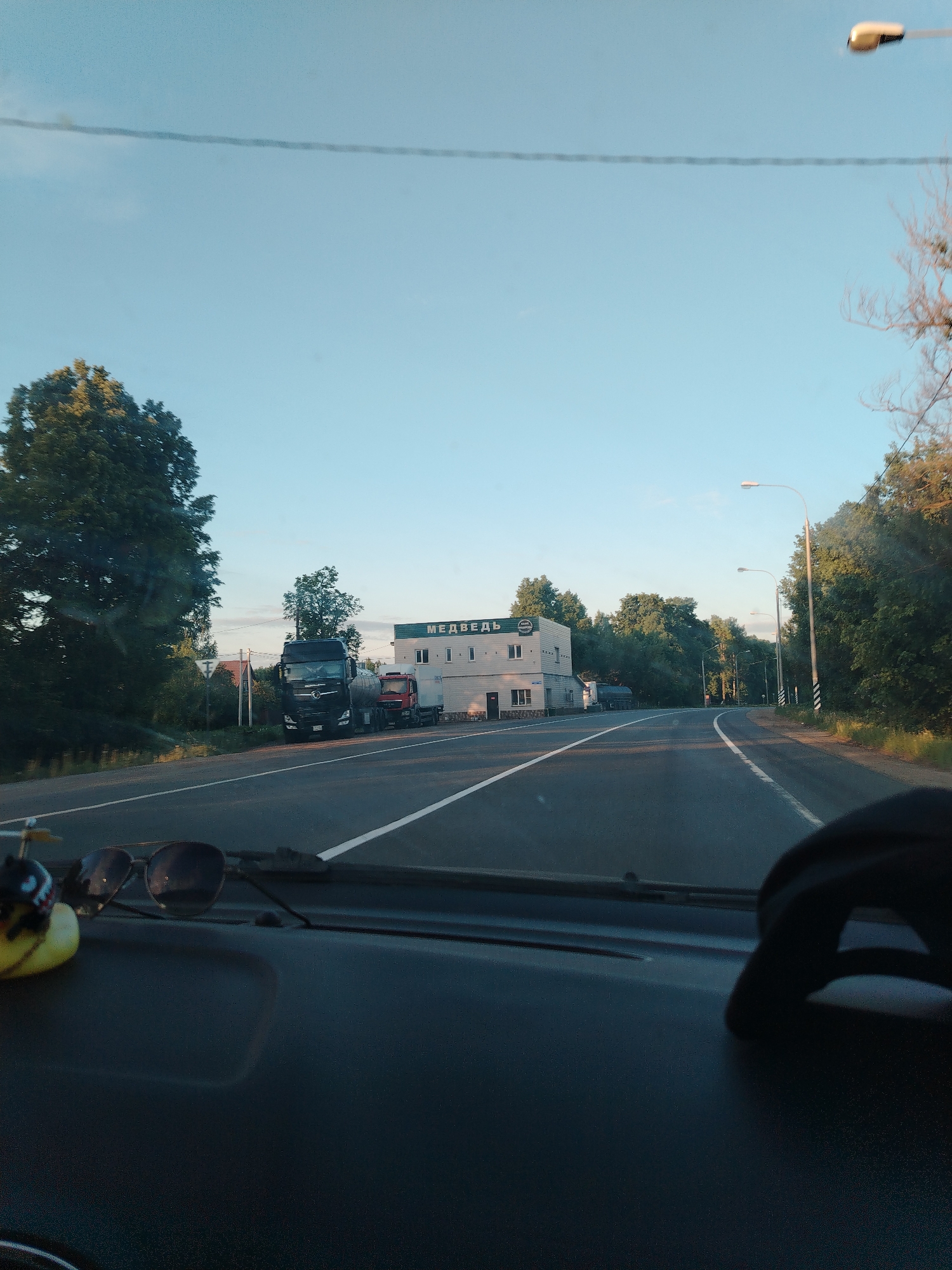 The image shows a view from inside a car driving on a road in Глебовское, Russia. The road is paved and has a white line marking the center. On the left side of the road, there's a building with a sign that says "МЕДВЕДЬ" (Bear). There are two trucks parked next to the building. Trees line both sides of the road. Streetlights are visible along the roadside. The sky is clear. A pair of sunglasses and a small toy are visible on the car's dashboard.
