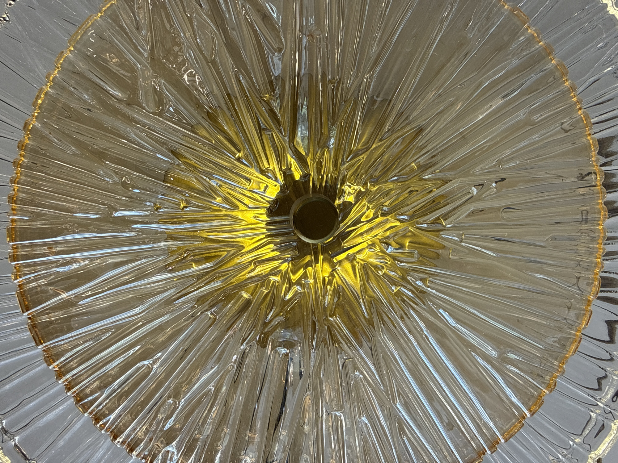 An overhead shot shows a close-up of a sunburst pattern glass fixture, possibly a light fixture or a decorative plate. The glass is clear with a warm amber hue towards the center. The pattern consists of many thin, radiating lines that create a starburst effect, giving the impression of rays. In the very center, there is a small, dark circular opening, suggesting a point of attachment or a light source. The lighting creates highlights and shadows that emphasize the texture and depth of the glass. The overall impression is one of intricate design and radiant light. The image is taken from an unknown angle within an unknown city.