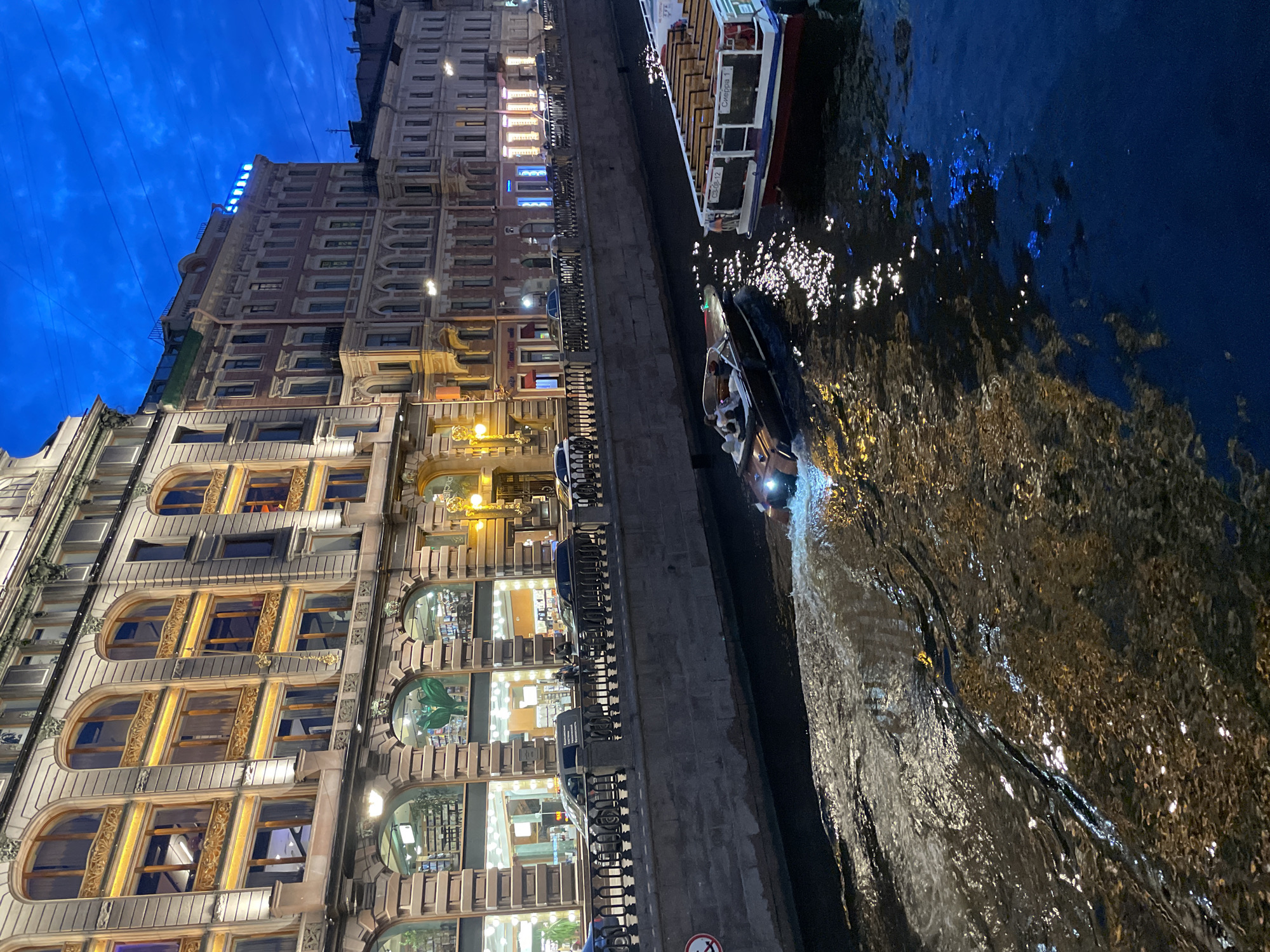 This is a vertical, eye-level, full shot of a canal with a speeding motorboat in the foreground. The water is dark blue and choppy, with reflections of building lights creating streaks of yellow and white. The motorboat is dark brown with white accents and has two people visible in it, a man and a woman. The motorboat is creating a white wake behind it.
To the left of the canal is a stone embankment with a black metal fence. Behind the fence is a building with a light beige facade and arched windows. The windows are illuminated from within, revealing a library or bookstore with shelves of books and a large green plant in one of the windows. Cars are parked along the street behind the fence.
To the right of the canal is another building with a pinkish-red facade and many windows. The building is illuminated, and there are lights visible on the roof. Further down the canal, a larger tour boat with a white roof and red accents is docked. The tour boat has the number "030-12" and the word "Сиерра 1" visible on its side. The sky is a deep twilight blue with some clouds. The city appears to be St. Petersburg, Russia.