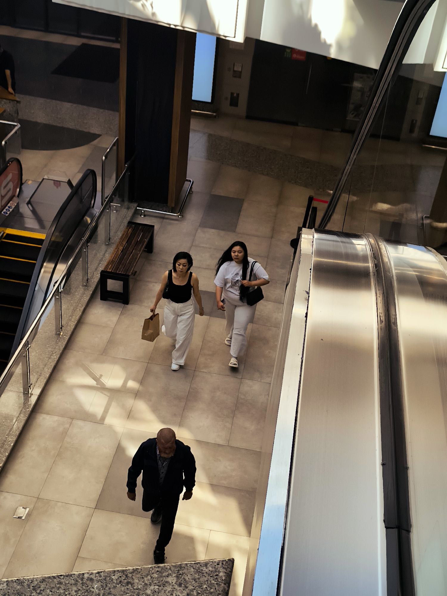An indoor scene shows a man and two women walking on a tiled floor, with an escalator visible to the right. The man, in the foreground, wears a dark jacket, a plaid shirt, and dark pants. He is walking away from the viewer. To his left, an escalator with yellow steps is partially visible. Further into the scene, two women are walking towards the viewer. The woman on the left wears a black tank top and white pants, carrying a brown paper bag. The woman on the right wears a white t-shirt and light-colored pants. A wooden bench is positioned between the women and the escalator. The environment appears to be a public space, possibly a mall or a transit station, with modern architecture and lighting.