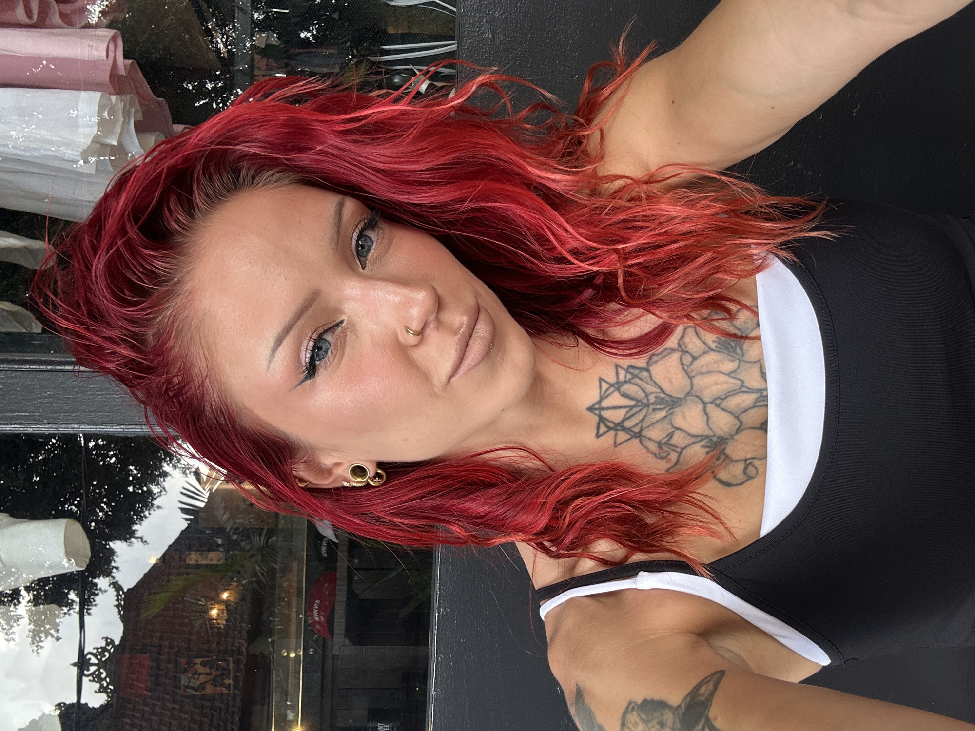 The image is a portrait of a woman with bright red hair, blue eyes, and nose and ear piercings. She is wearing a black tank top with a white underlayer. She has tattoos on her left arm and chest. The background appears to be a reflection in a window, showing a building with traditional Indonesian architecture and lush greenery. The woman is looking directly at the camera with a neutral expression. The setting appears to be outdoors, possibly a cafe or shop in North Kuta, Indonesia.