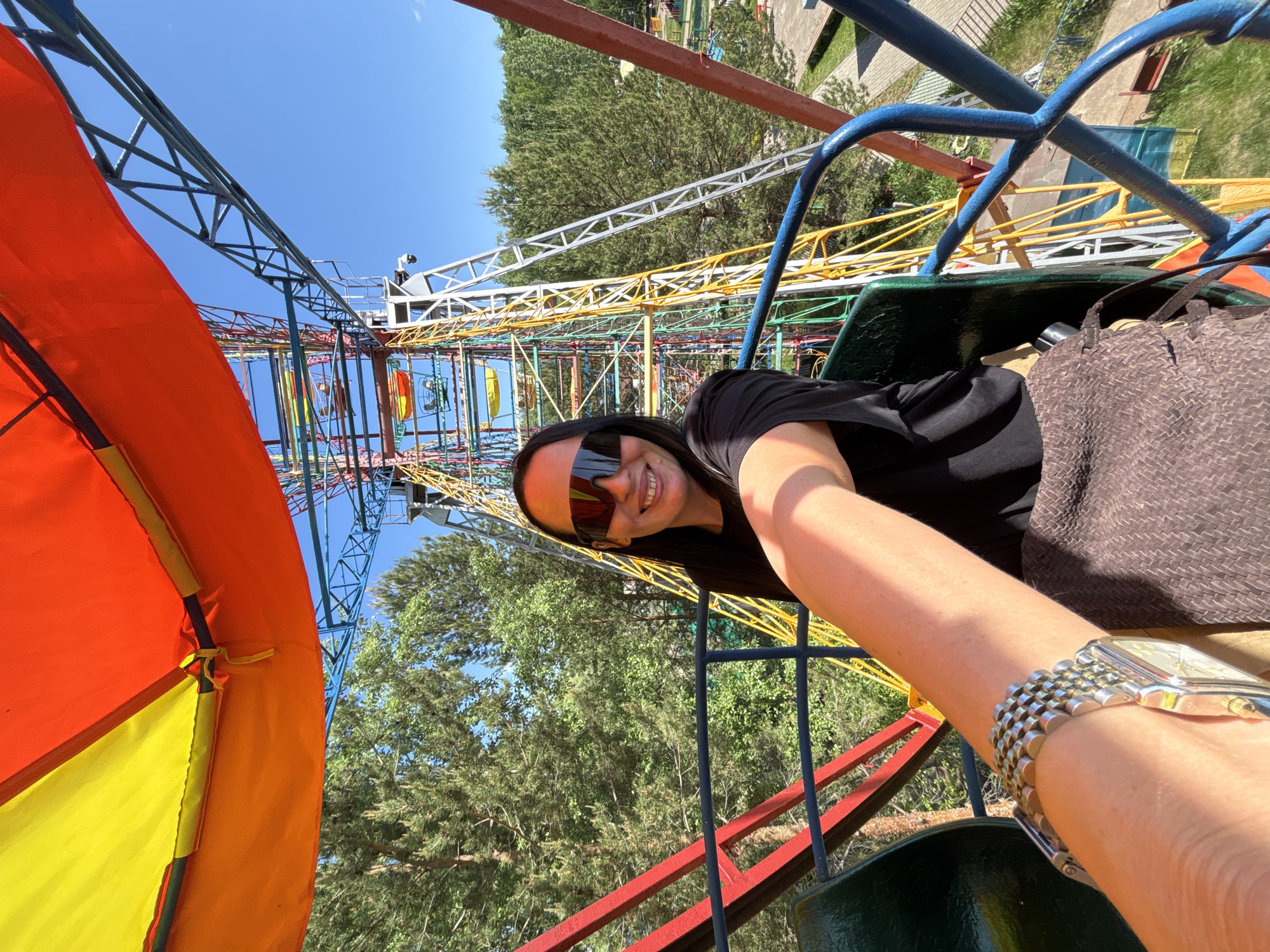 The image shows a person in a small Ferris wheel, possibly in an amusement park. The person is wearing a black t-shirt and sunglasses, and they are smiling at the camera. The Ferris wheel is painted in bright colors, and the background includes trees and a clear blue sky. The location may be in Chaikovsky, Russia, although there is no verifiable information to confirm that. The person is taking a selfie from the Ferris wheel. They are wearing a silver watch.