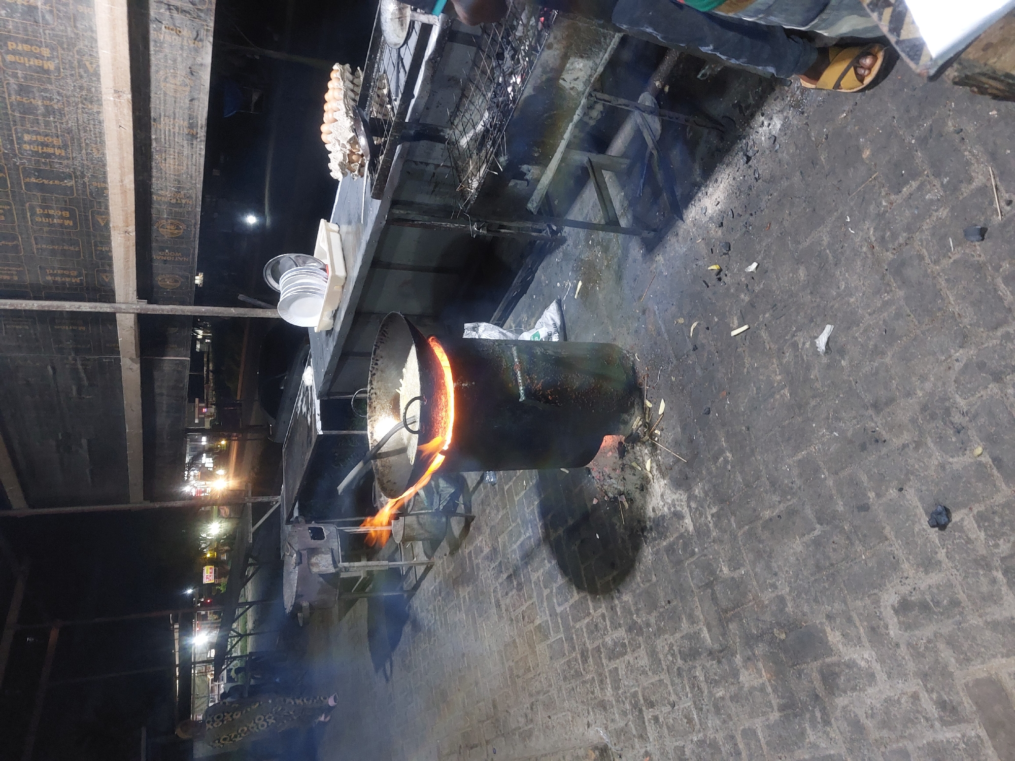 This image shows a street food vendor's setup at night. A large metal container with an orange glow emanating from it, likely a cooking device or oven, sits on a metal stand. Smoke and flames can be seen near the container. To the left, a shelf holds a carton of eggs and a stack of disposable bowls or containers. A metal grill is positioned above the container. A person is visible, wearing sandals, standing near the setup, possibly attending to the cooking. The scene is illuminated by streetlights in the background, suggesting an urban setting, identified as Dar es-Salaam, Tanzania. The ground is paved.