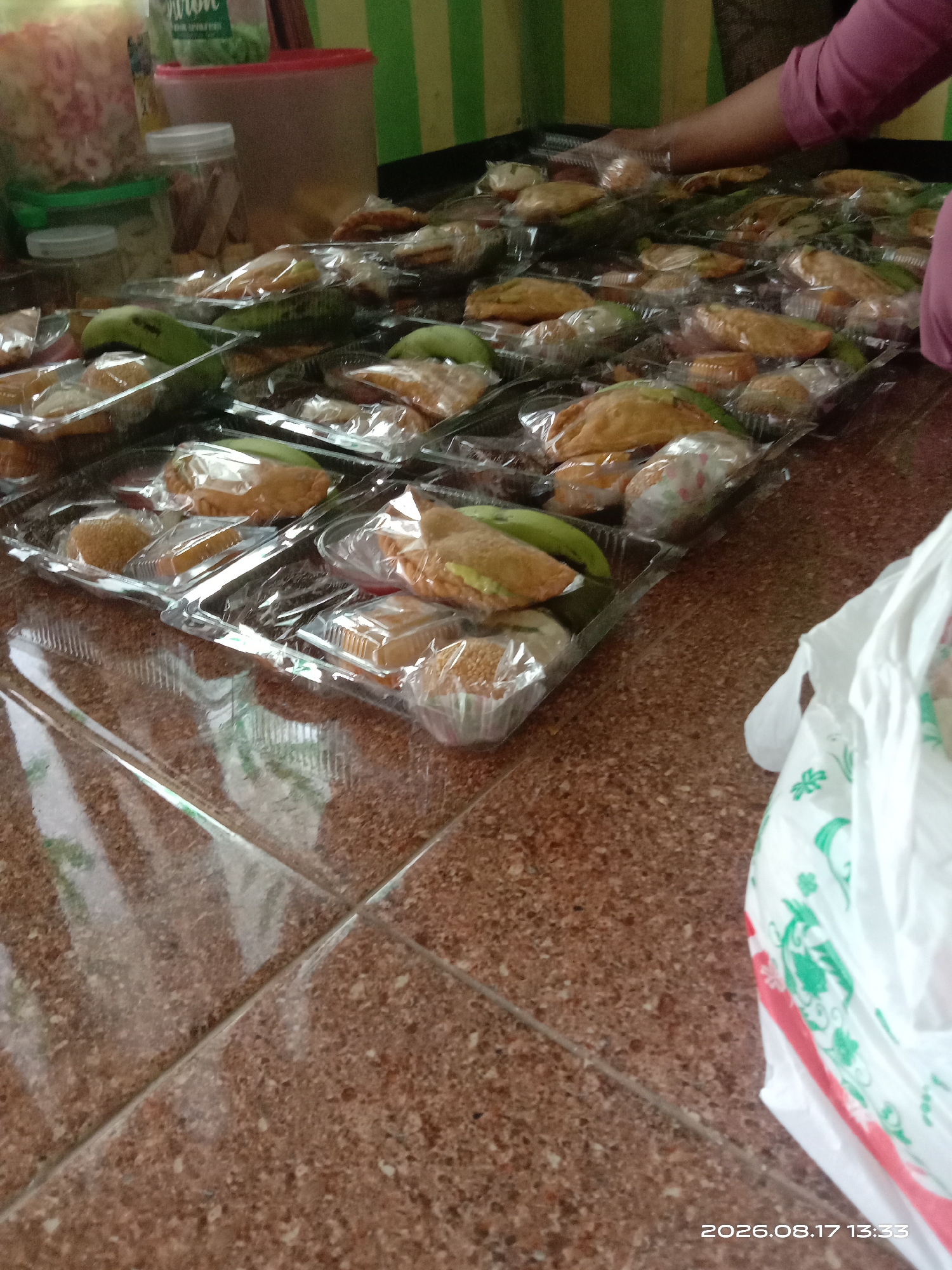 The image depicts a street-level view of a food stall or a vendor preparing food for sale in Banyuarang, Indonesia. In the foreground, a portion of a brown tiled floor is visible, with a white plastic bag adorned with green floral patterns and red accents on the right side. The bag appears to be partially filled. In the mid-ground, a long table is laden with numerous pre-packaged food items, arranged in clear plastic containers. These items include what appear to be pastries or snacks, some with a golden-brown fried appearance and others with a green filling, possibly fruit or a savory paste. Some of the packages also contain small, round, bread-like items coated in sesame seeds. A hand, presumably of a vendor, is partially visible on the right side of the frame, wearing a pinkish-purple shirt and reaching towards the food containers. In the background, out of focus, are various containers, including a large red-lidded container and a green plastic container filled with what looks like snack items. There are also hints of vertical green and yellow striped fabric, possibly a backdrop or awning. The lighting suggests an indoor or shaded outdoor setting. The overall impression is one of food preparation and sale, likely for a local market or event.