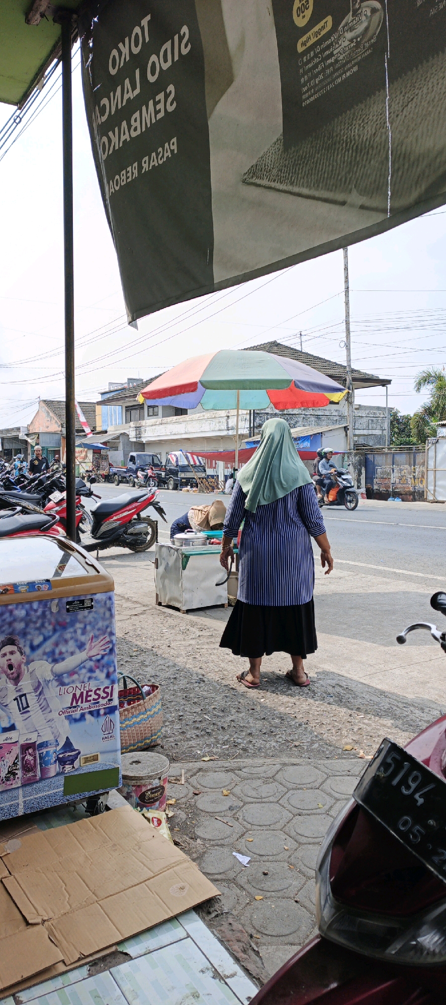 This image depicts a street scene in Kasiyan, Indonesia, likely near a market or roadside stalls. In the foreground, a large refrigerator adorned with a picture of Lionel Messi is visible, suggesting it might be used for selling beverages. A woven basket sits beside it, along with scattered cardboard and debris on the ground. A woman wearing a light green hijab and a striped blue and white dress stands facing away from the camera, her hands by her sides. She is wearing sandals. Behind her, a colorful umbrella provides shade over a food stall, where a person is bent over. Various motorcycles are parked in the background, and a few vehicles, including a small truck, are on the road. Buildings line the street, some appearing to be shops with awnings. Overhead, power lines crisscross the sky. The overall impression is one of a bustling, everyday street market environment.