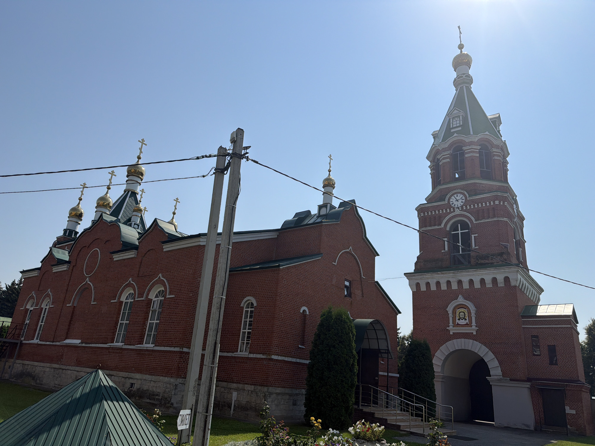 The image depicts the Church of the Assumption of the Blessed Virgin Mary in Troekurovo, Russia, bathed in bright sunlight under a clear blue sky. The primary subject is the red brick church, characterized by its golden domes topped with crosses and a tall, square bell tower. The church features arched windows and ornate architectural details. In the foreground, a conical green metal structure is visible, along with a gray utility pole with multiple wires. The overall environment appears to be a well-maintained church complex with some greenery and flowers. There are no people or animals visible in the frame.