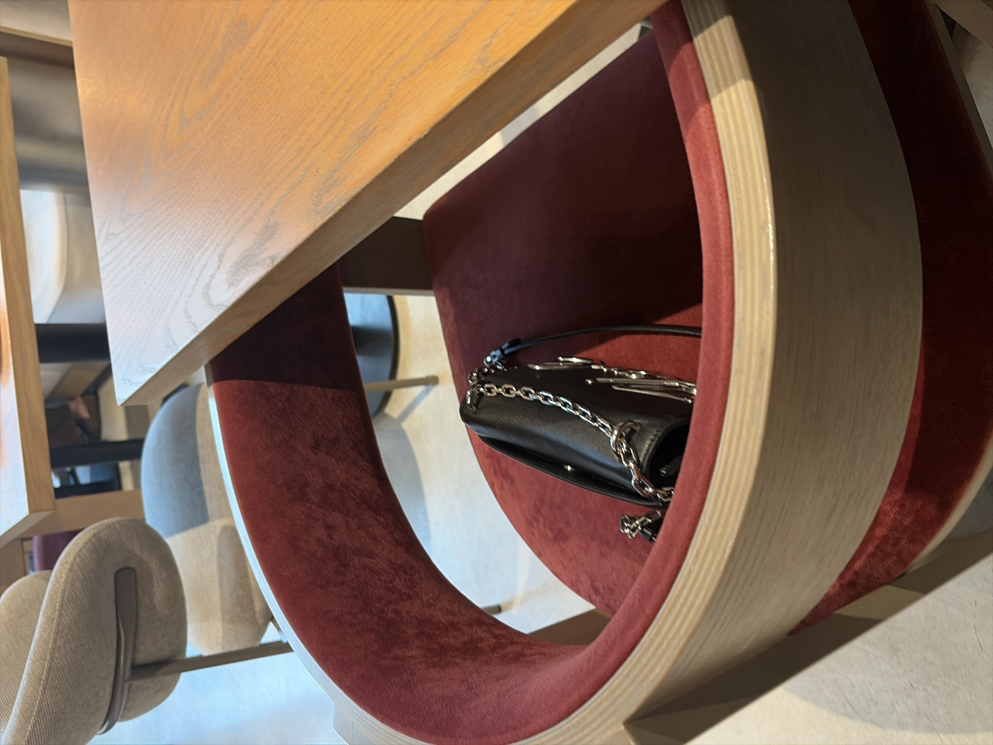 The image shows a close-up of a red upholstered chair with a curved wooden frame. A black leather handbag with a silver chain strap is placed on the seat of the chair. The chair is positioned at a table, and the wooden surface of the table is visible in the upper right corner of the image. The background is blurred, but hints of other chairs and tables suggest an indoor setting, possibly a cafe or restaurant. There are no people visible in the image. The lighting suggests an indoor environment, possibly with natural light coming from a window. The overall impression is one of a still life, focusing on the details of the chair and the handbag.