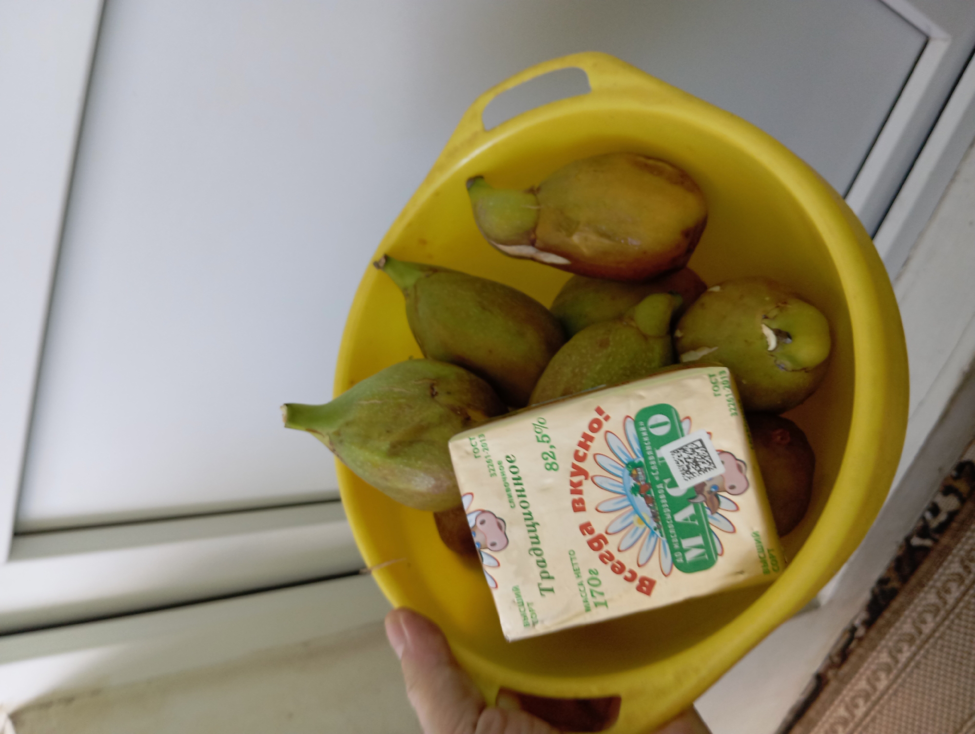 A hand is holding a yellow plastic bowl filled with several green and yellow figs. A package of butter, labeled in Russian, is also in the bowl, partially obscuring some of the figs. The butter package features a cartoon cow and text that translates to "Traditional" and "Always tasty!". The background appears to be indoors, possibly near a window or door with a light-colored wall and trim. The scene does not contain any elements that would indicate its location in Temryuk, Russia.