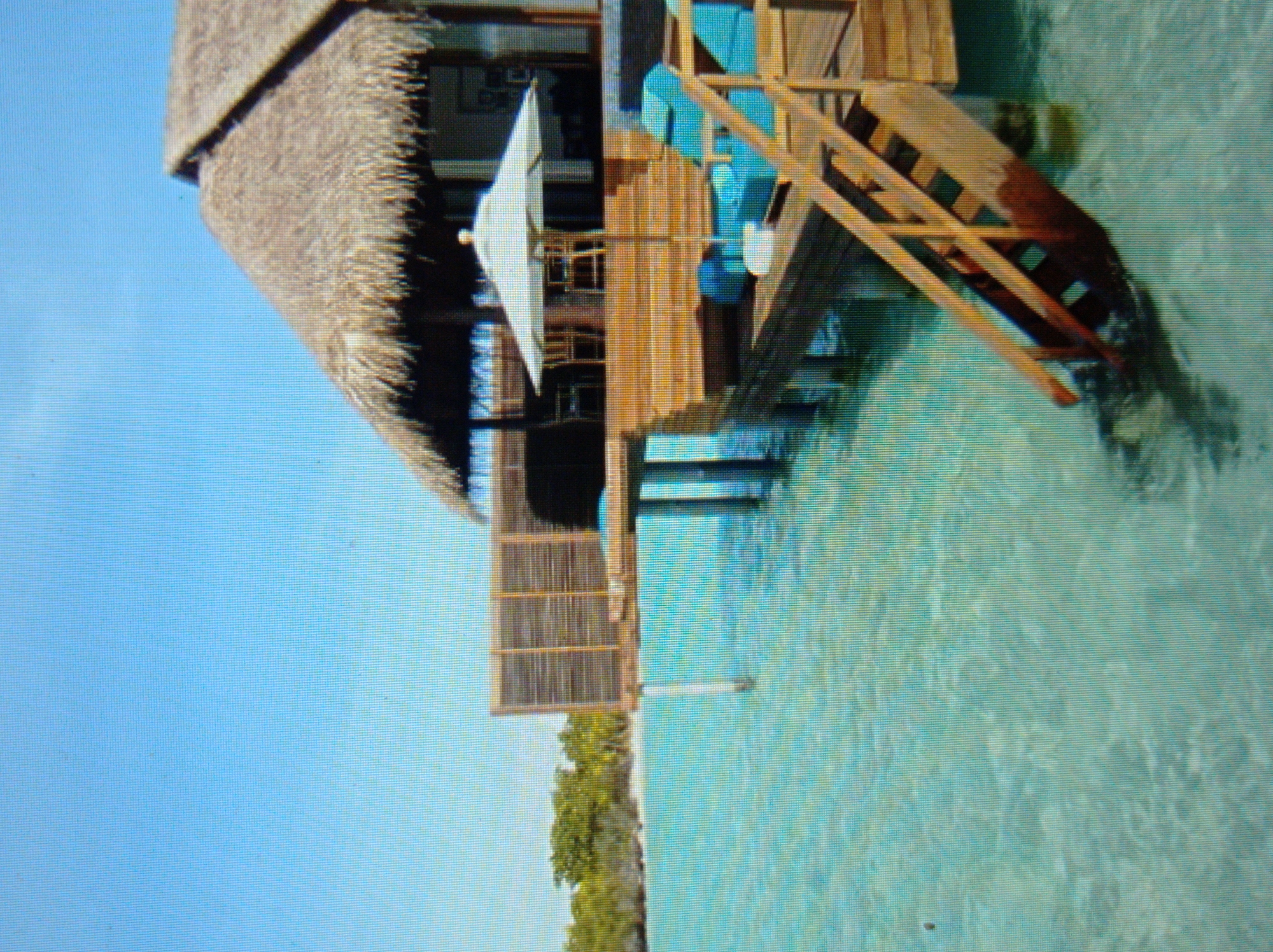 The image displays a tropical resort scene. In the foreground, turquoise water laps against a wooden deck with a set of stairs leading into the water. On the deck, there are two blue lounge chairs and a white umbrella. Behind the deck, a thatched-roof structure with wooden walls and furniture is visible. In the background, lush green vegetation borders the water. There are no people visible in the image. The scene is set on a bright, sunny day with a clear blue sky. There is no indication that this scene is located in the city of Mukachevo, Ukraine.