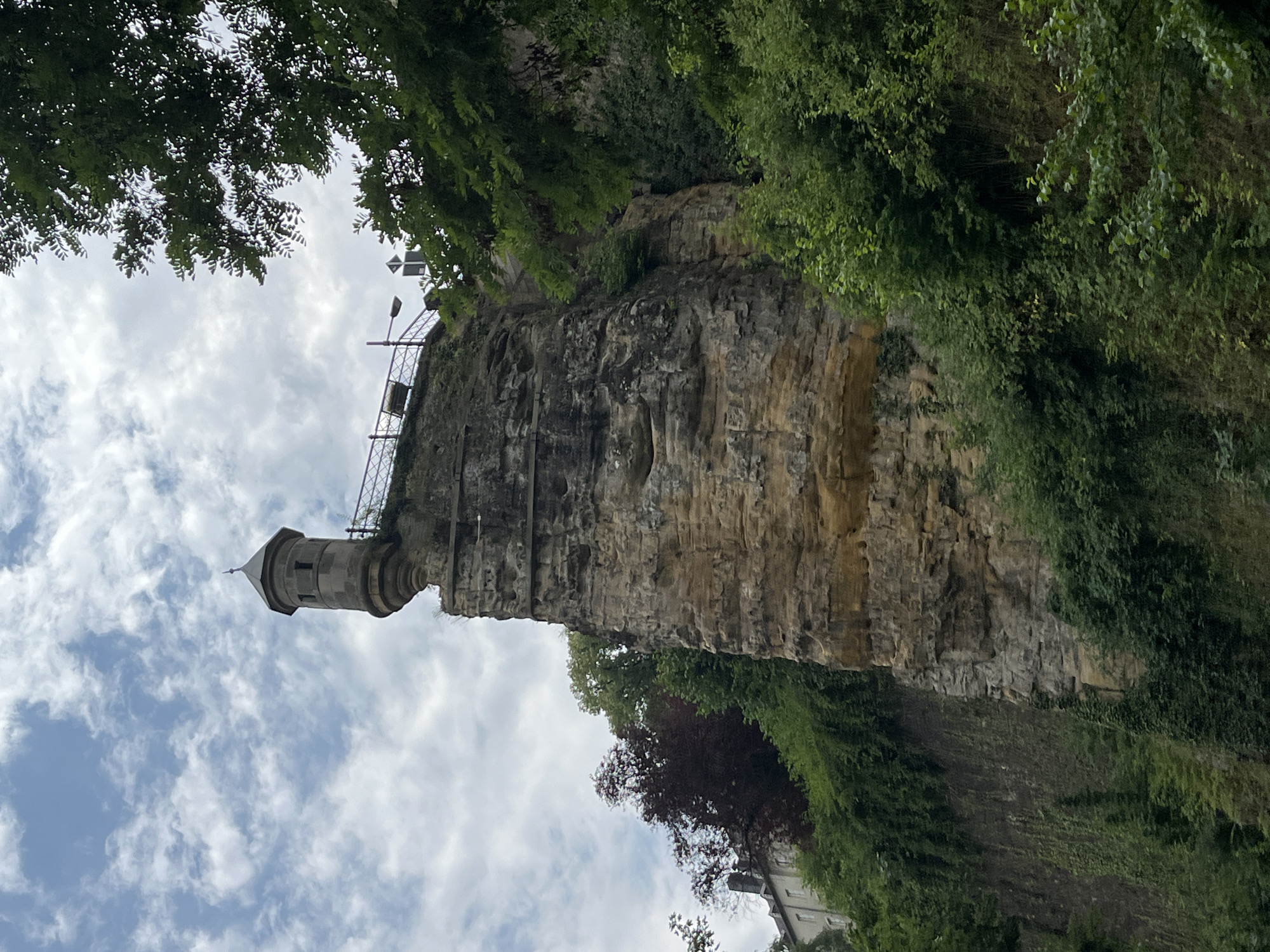 A stone tower with a pointed roof stands on a rocky cliff. The tower is cylindrical with a wide base and is decorated with moldings. A metal railing surrounds the top of the cliff. Below the cliff, lush green trees and vegetation grow, with a few buildings visible in the distance. The sky is cloudy, with patches of blue visible.