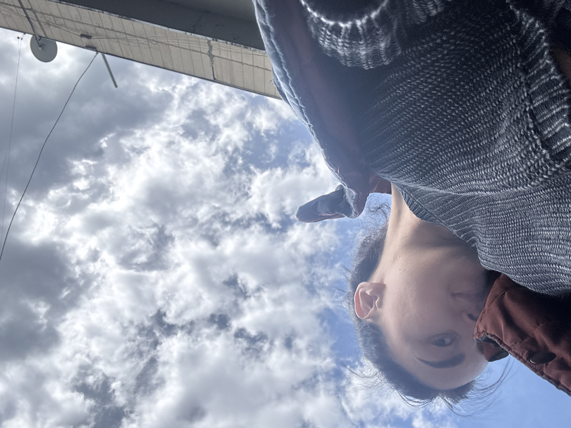 The image captures a unique low-angle perspective, primarily featuring a person and a cloudy sky. The subject, a person, is visible on the right side of the frame, looking downwards towards the camera. They have dark hair, with part of their face, including an ear and one eye, clearly visible. The person is wearing a dark, textured sweater over a reddish-brown jacket or vest.
In the upper left portion of the image, an overhead structure is partially seen. It appears to be a light-colored canopy, awning, or part of a building's underside, with visible lines suggesting a textured surface or support framework. A small, circular, dish-like object and some thin wires are suspended from this structure.
The majority of the background is filled with a bright, partly cloudy sky. Patches of blue sky are visible between various shades of white and gray clouds. The overall lighting suggests it is daytime. The unusual angle, with the person's head rotated towards the right side, indicates the photo was likely taken from a very low vantage point, possibly a selfie. The person's expression is neutral. There is no visible text in the image.