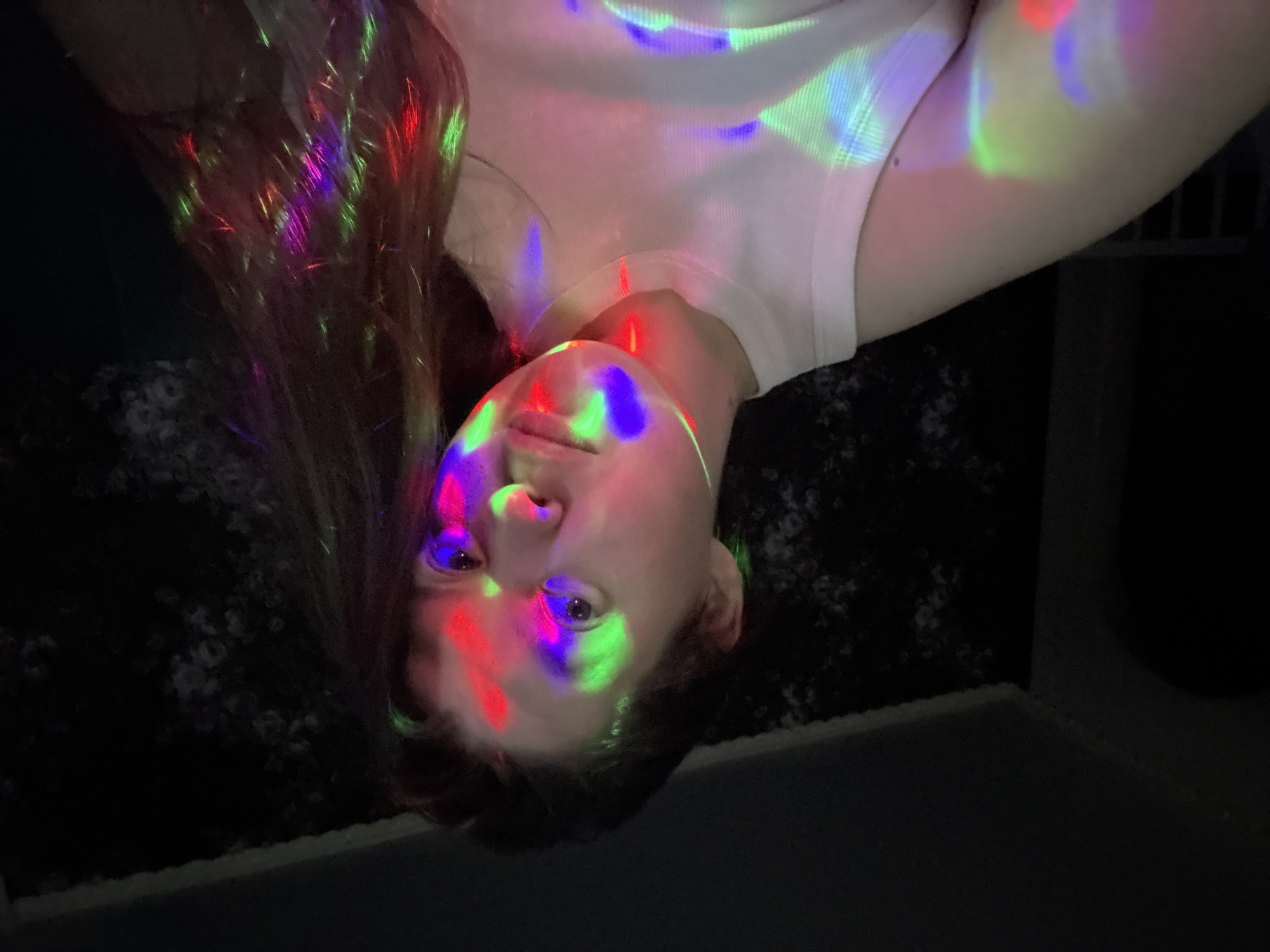 A person with fair skin and light brown hair is lying down, looking up at the camera. Their face is illuminated with red, green, and blue lights. They are wearing a white top. The background appears to be a dark room with a textured surface. There is no definitive evidence to confirm that the image was taken in Марковский, Russia.