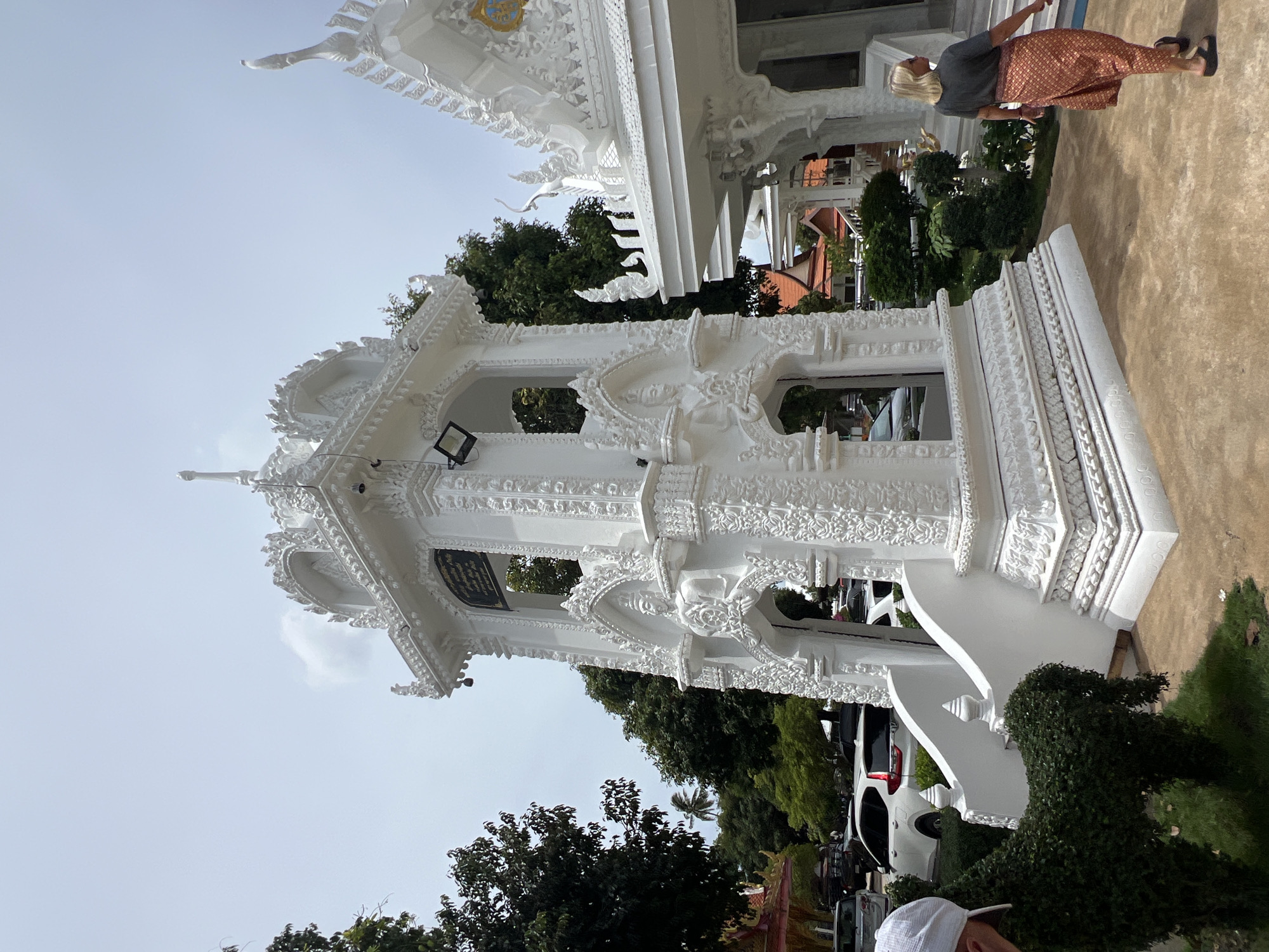In Baan Bo Phut, Thailand, a white, ornate bell tower stands prominently in front of a white temple. The tower features intricate carvings and arched openings, with a seated figure visible within one of the lower arches. A person, identified as a woman with blonde hair wearing a gray t-shirt and a patterned skirt, walks away from the camera towards the right side of the frame, near the temple structure. To the left of the bell tower, a portion of a white cap is visible on a person who is mostly out of frame. In the foreground, a topiary shaped like a horse is situated on a patch of grass. The background shows trees, parked cars, and other buildings, indicating an outdoor, public setting, possibly a temple complex. The sky is overcast.