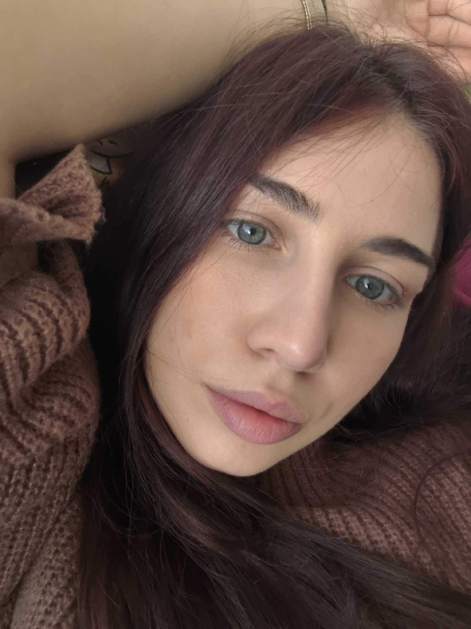 A close-up selfie shows a woman with long, auburn hair, light skin, and blue eyes. She is wearing a brown knitted sweater. The background is blurry, obscuring any identifiable location in Stara Zagora, Bulgaria. No actions or interactions are discernible.