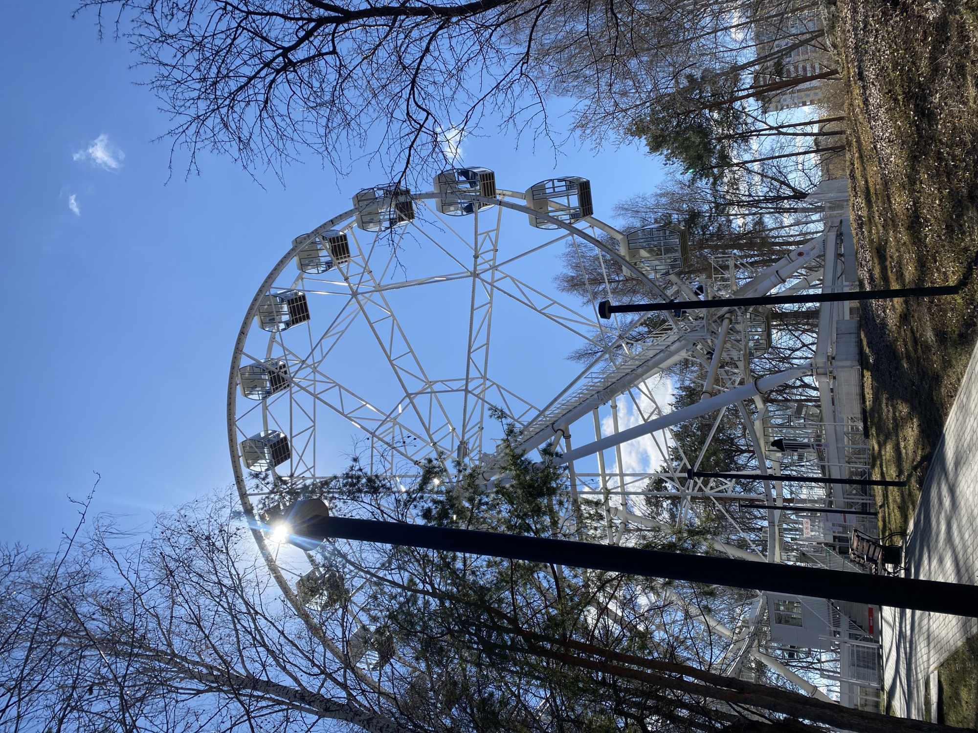 The image features a Ferris wheel in what appears to be a park. The wheel is white with enclosed gondolas. It is partially obscured by bare trees in the foreground, with the sun visible behind one of the trees. The sky is blue with a few clouds. In the background, there are additional trees and a building. The location is Yekaterinburg, Russia. A lamppost is visible near the bottom of the frame. A walkway with benches is present to the right of the Ferris wheel.