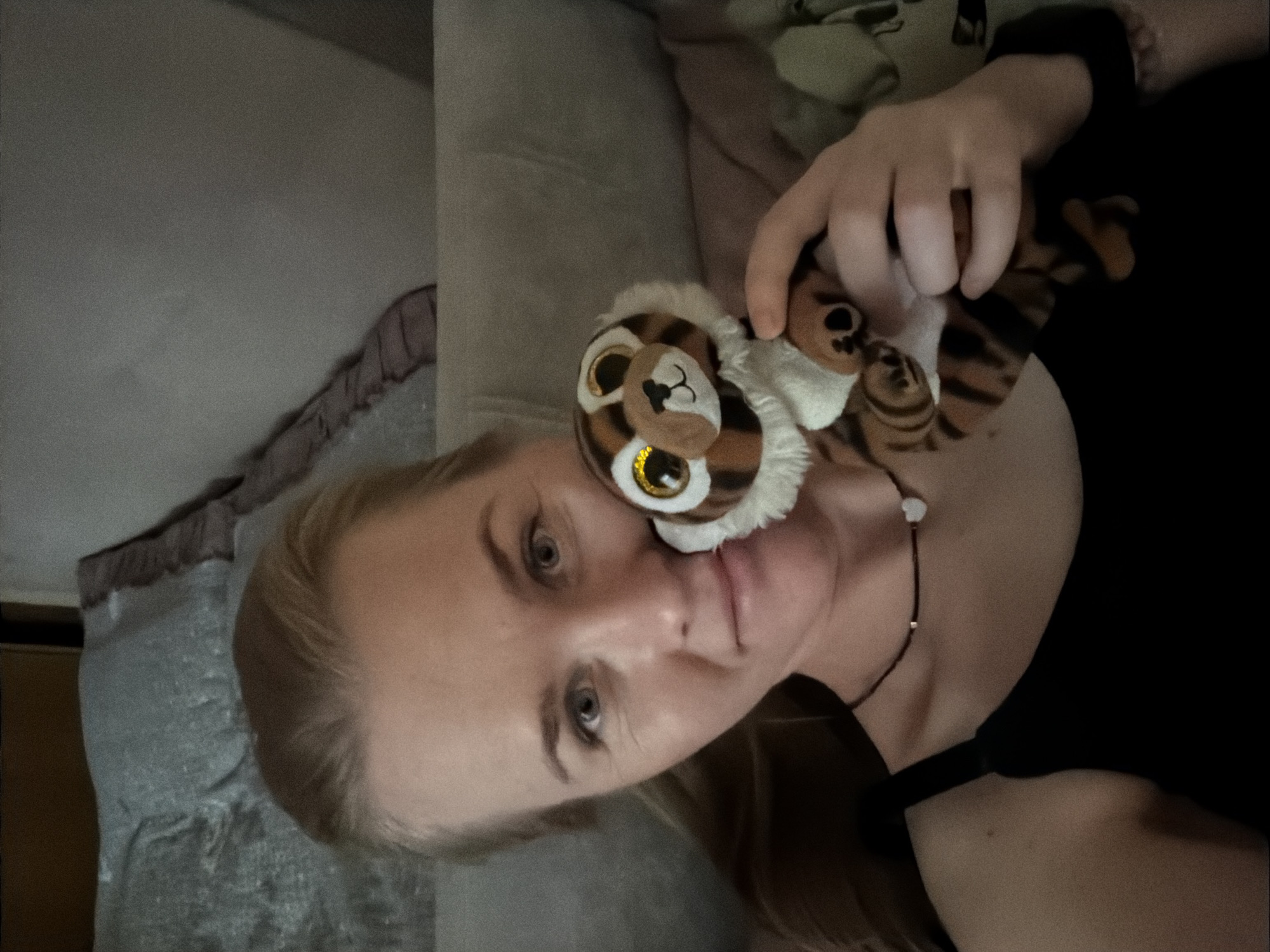 A woman with blonde hair, wearing a black tank top and a necklace with a heart pendant, lies on a bed, holding a stuffed tiger. The background shows a gray pillow and a portion of a wall or headboard. The image appears to be a selfie taken indoors. The location is not specified and cannot be determined from the image.