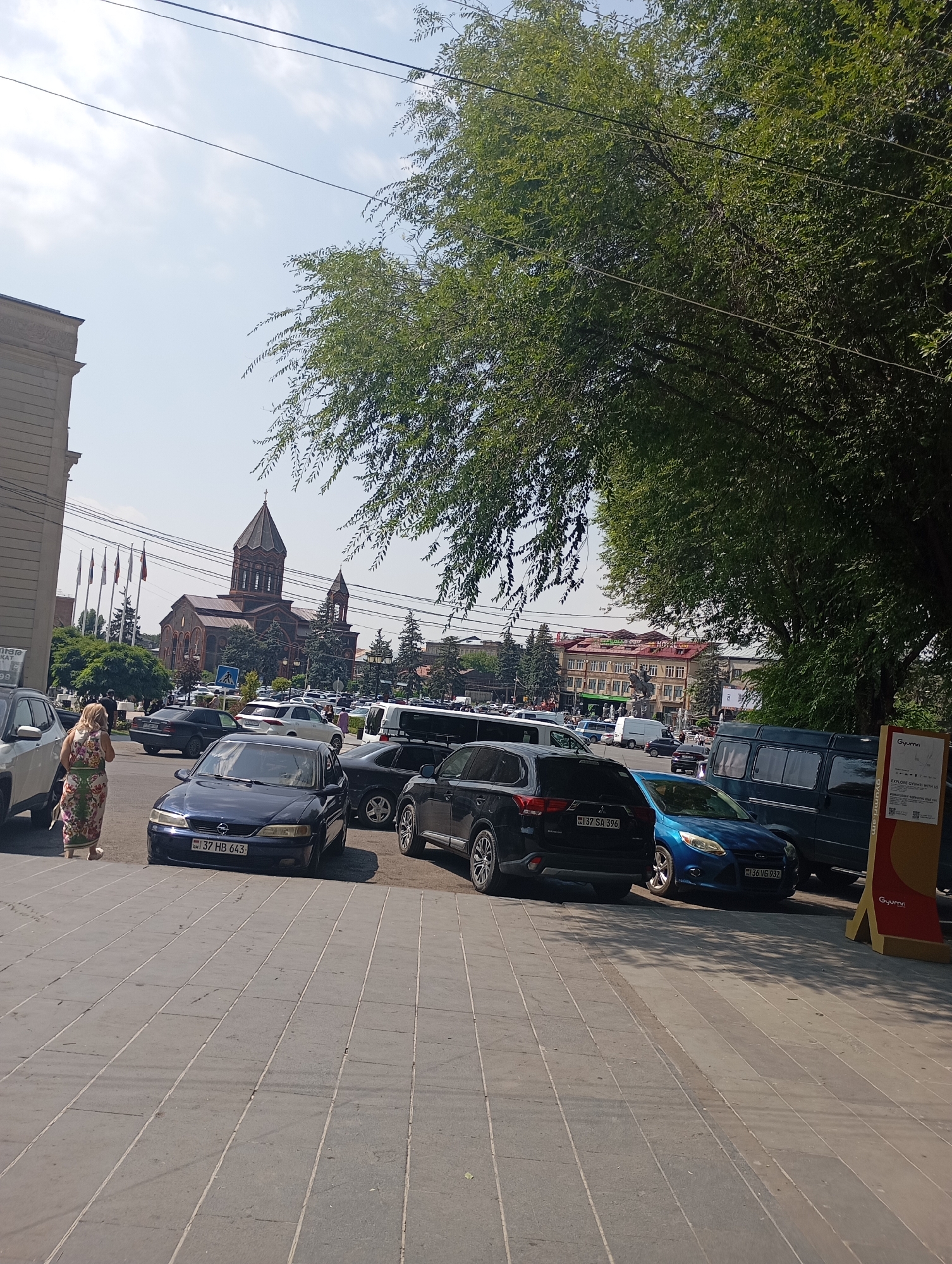The image depicts a street scene in Gyumri, Armenia, with a prominent church in the background, characterized by its dark stone construction and a multi-tiered roof topped with a cross. The church has a bell tower and arched windows. In the foreground, several cars are parked on the street, including a dark blue sedan with a license plate that reads "37 HB 643", a black SUV, and a blue hatchback. A woman in a floral dress is walking away from the camera on a paved sidewalk. The street is lined with trees, and buildings with multiple stories and balconies are visible across the street from the church. The sky is clear and blue, with some power lines visible. A yellow stand with signage is partially visible on the right side of the image.