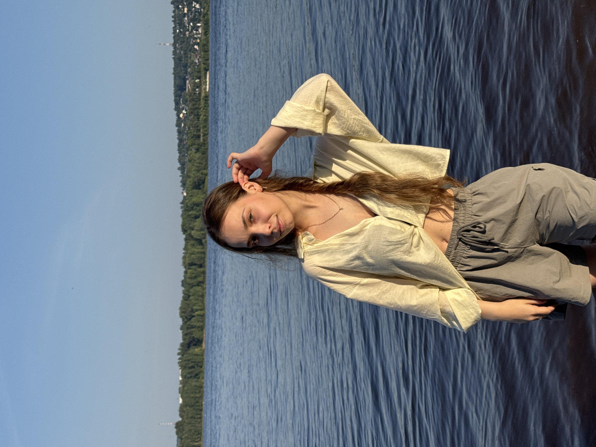 A young woman with long, braided brown hair stands partially submerged in calm blue water, looking towards the camera. She is wearing a light yellow, oversized, unbuttoned shirt that is tied at the waist, exposing her midriff, and grey drawstring shorts. Her right hand is raised to her ear, and she is wearing a thin gold necklace and small hoop earrings. The background shows a wide body of water with a dense line of green trees on the opposite bank, beyond which are houses and buildings, characteristic of the city of Noviy, Russia. The sky is clear and blue.