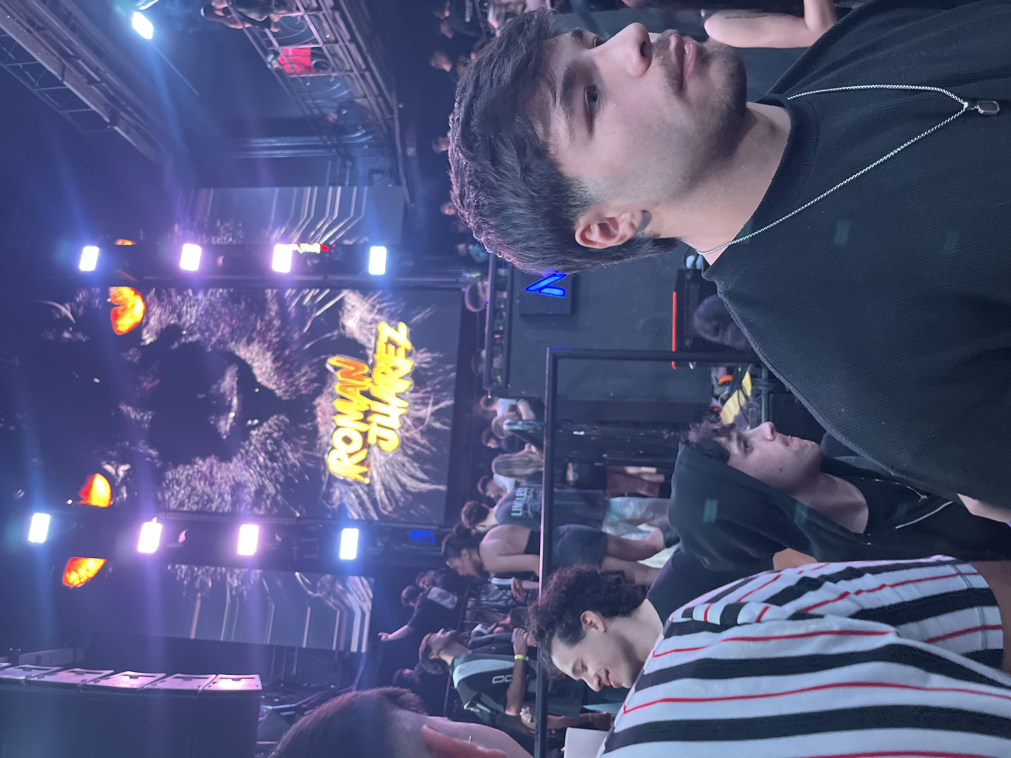 The image shows an indoor event. A crowd of people are seated or standing, some looking towards a stage. On the stage, a large screen displays an image of a wolf and the text "ROMAN JUAREZ". Lighting equipment illuminates the stage. In the foreground, several people are partially visible. One person is wearing a black long sleeve shirt and a silver chain necklace. Another is wearing a shirt with black and white stripes.
The information about the location in Buenos Aires, Argentina is not detectable.