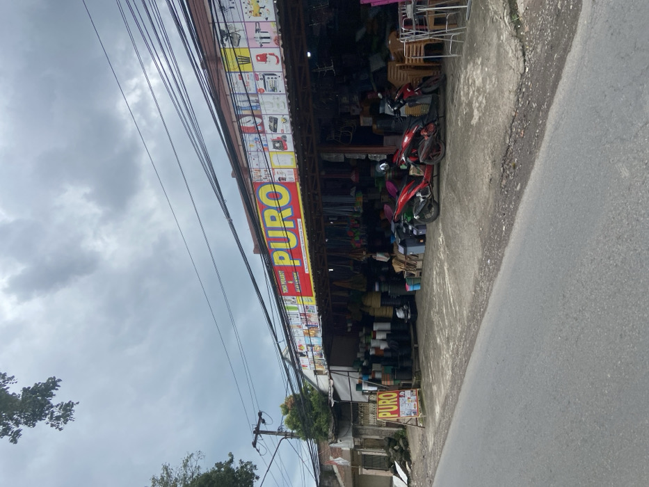 The image shows a store front in Ciparay, Indonesia, located at latitude: -7.022, longitude: 107.679. The store front is labeled "TOKO PERALATAN PURO," and the word "PURO" is displayed prominently in large, yellow, block letters on a red background. The store front is lined with stacks of various household goods, including buckets, mops, brooms, and chairs. Two red motorcycles are parked in front of the store, and one is facing the viewer. The store is located on the right side of the image, facing a paved road. The road is empty, and the environment includes a cloudy sky, power lines, and a lush green tree on the left side of the frame.