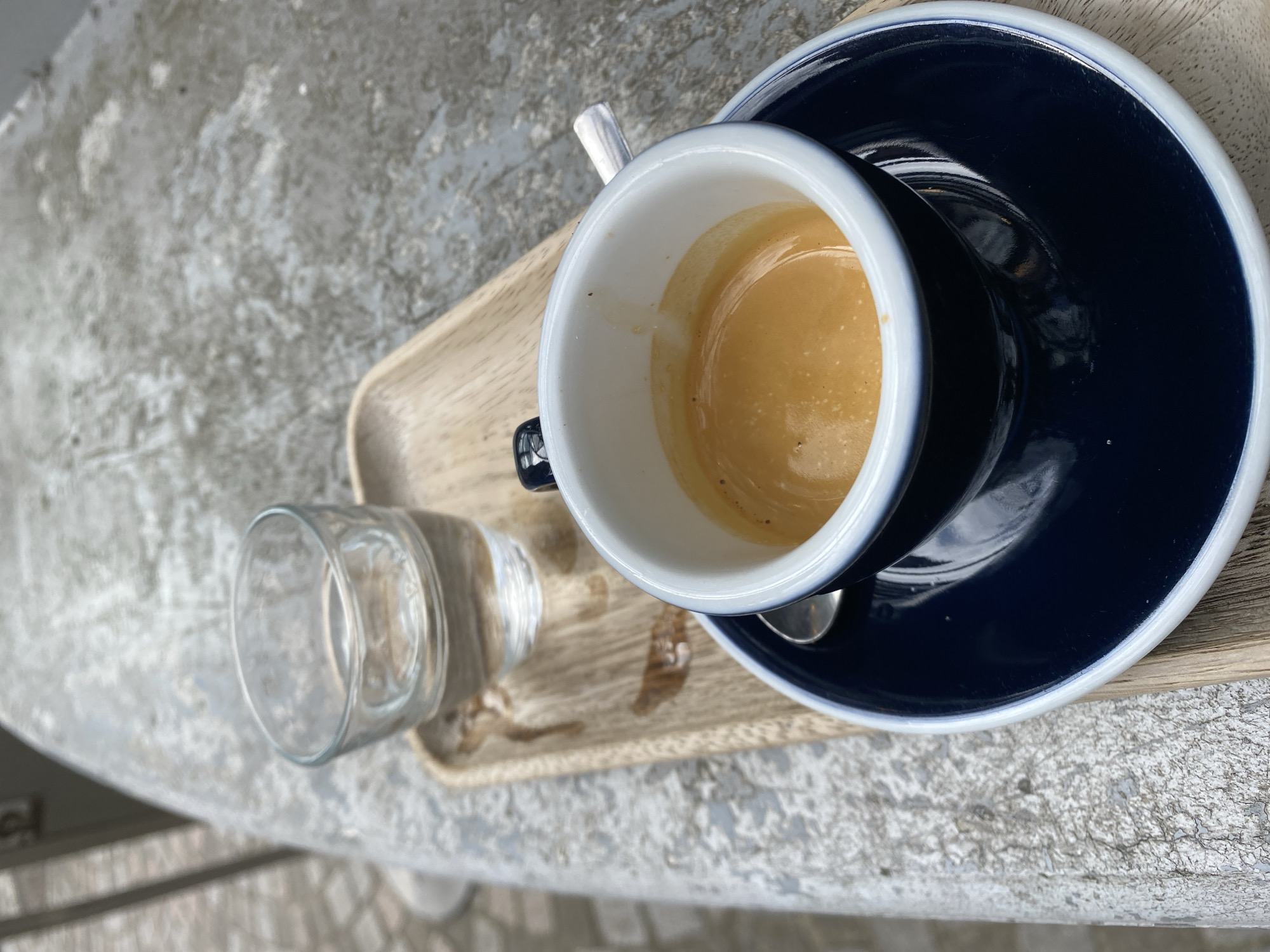 The scene is a close-up of a wooden tray on a grey textured surface. The tray holds a dark blue coffee cup with a white interior, containing a partially consumed espresso. A matching blue saucer sits beneath the cup, and a small silver spoon is nestled against the cup. A glass of water is positioned beside the cup on the tray. The location may be Strasbourg, France, based on the architectural styles and common cafe culture of the region, which can be seen in the background outside.