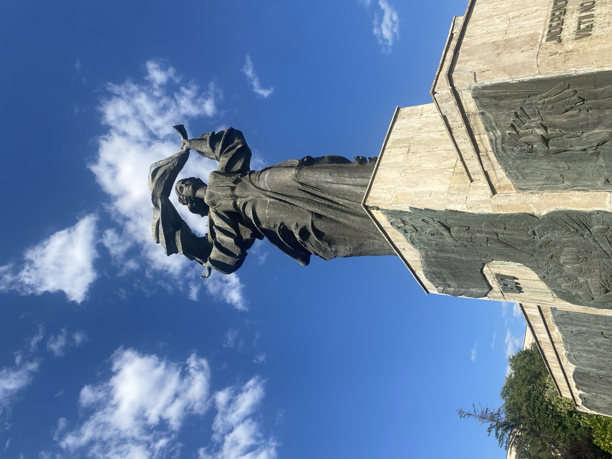 The image captures a monumental statue in the city of Iași, Romania, against a backdrop of a bright blue sky with scattered white clouds. The statue, predominantly dark in color, appears to be made of bronze or a similar metal and depicts a female figure holding a large, draped fabric aloft with both hands. The figure is positioned on a multi-tiered, light-colored stone pedestal, which is adorned with bas-relief carvings.
The lower part of the pedestal features extensive etched scenes that appear to depict historical or symbolic narratives. One section shows figures on horseback, possibly in a procession or battle. Another section displays the year "1917" in a distinct font. Towards the bottom right of the pedestal, visible text reads "INDEPENDENȚA" and partially visible text below it reads "VIETII NOASTRE", which translates to "Independence of our lives".
Green foliage and trees are visible to the left and behind the statue, suggesting a park or public space setting. The lighting indicates it is daytime, with sunlight casting shadows that highlight the texture and form of the statue and its carvings. The perspective is from a low angle, looking up at the grandeur of the monument.
