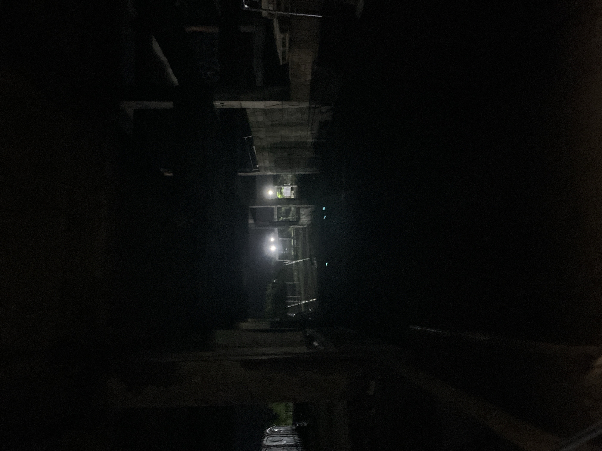 A dark, low-light, wide-angle shot captures an outdoor scene at night in the city of Poti, Georgia. The image appears to be taken from within an unfinished or abandoned structure, with concrete pillars and walls visible on the left and right sides of the frame. In the background, a row of portable toilets is lined up on the left, partially obscured by shadows. Further back, a pathway or road is illuminated by bright overhead lights, casting long shadows and highlighting the dark, flat ground. The overall atmosphere is dim and somewhat eerie, with limited visibility beyond the illuminated area.
