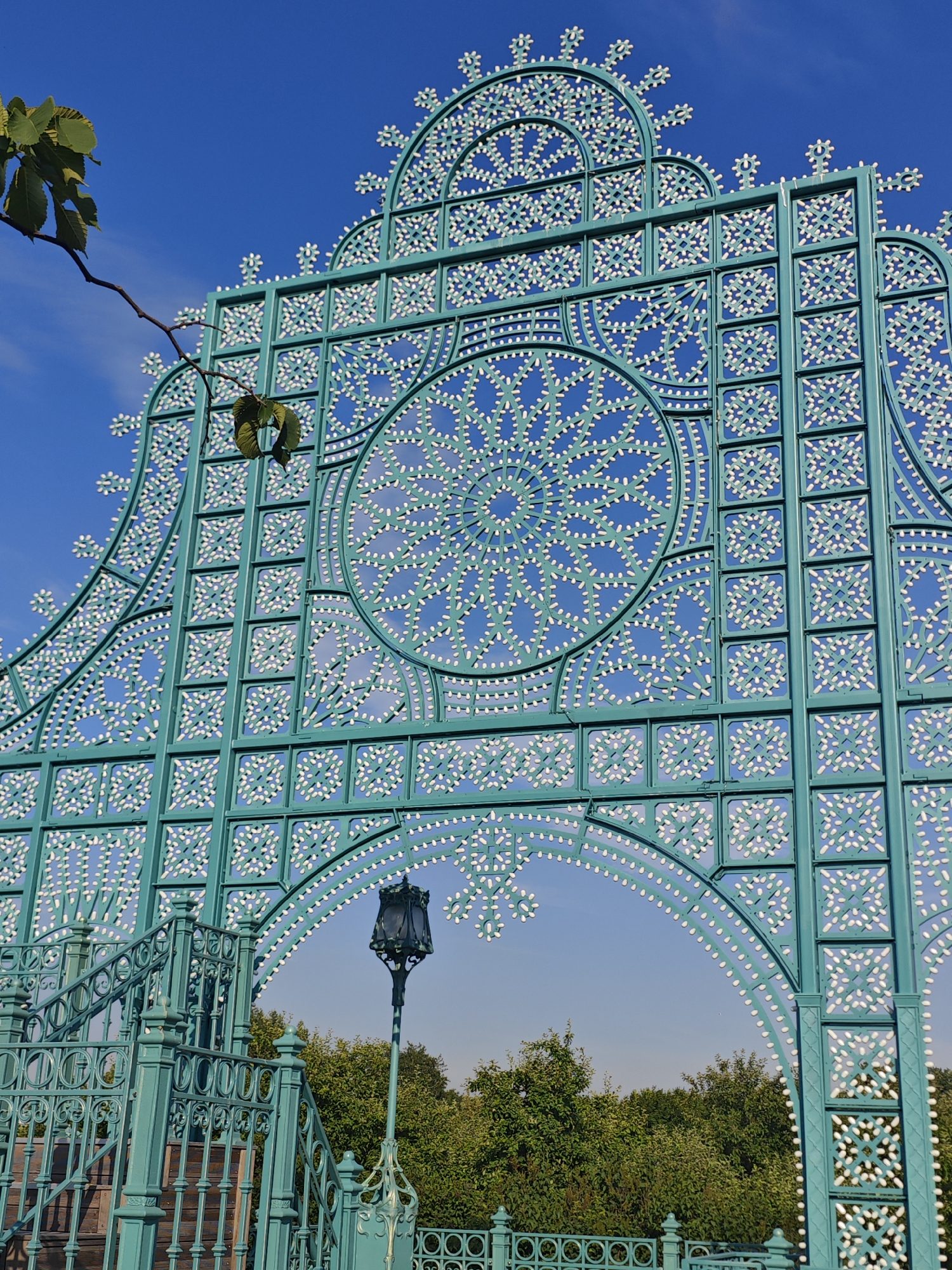 The image captures a portion of a teal-colored, ornate metal structure against a clear blue sky with a few wispy clouds. The structure features intricate geometric patterns with small white embellishments that resemble dots or pearls. A large circular motif with a starburst pattern is visible in the upper center of the structure. To the left, a branch with green leaves extends into the frame from the top left corner. In the lower portion of the image, a teal-colored staircase with decorative railings and wooden steps is visible. A vintage-style teal streetlamp stands to the right of the staircase. In the background, a dense line of green trees can be seen.
This structure is located in Gorky Park (Park Gorkogo) in Moscow, Russia. Specifically, it is part of the "Fountain Arch" (Fountannyi Ark), a prominent decorative element in the park, often illuminated at night. The arch serves as an entrance or visual focal point within the park.