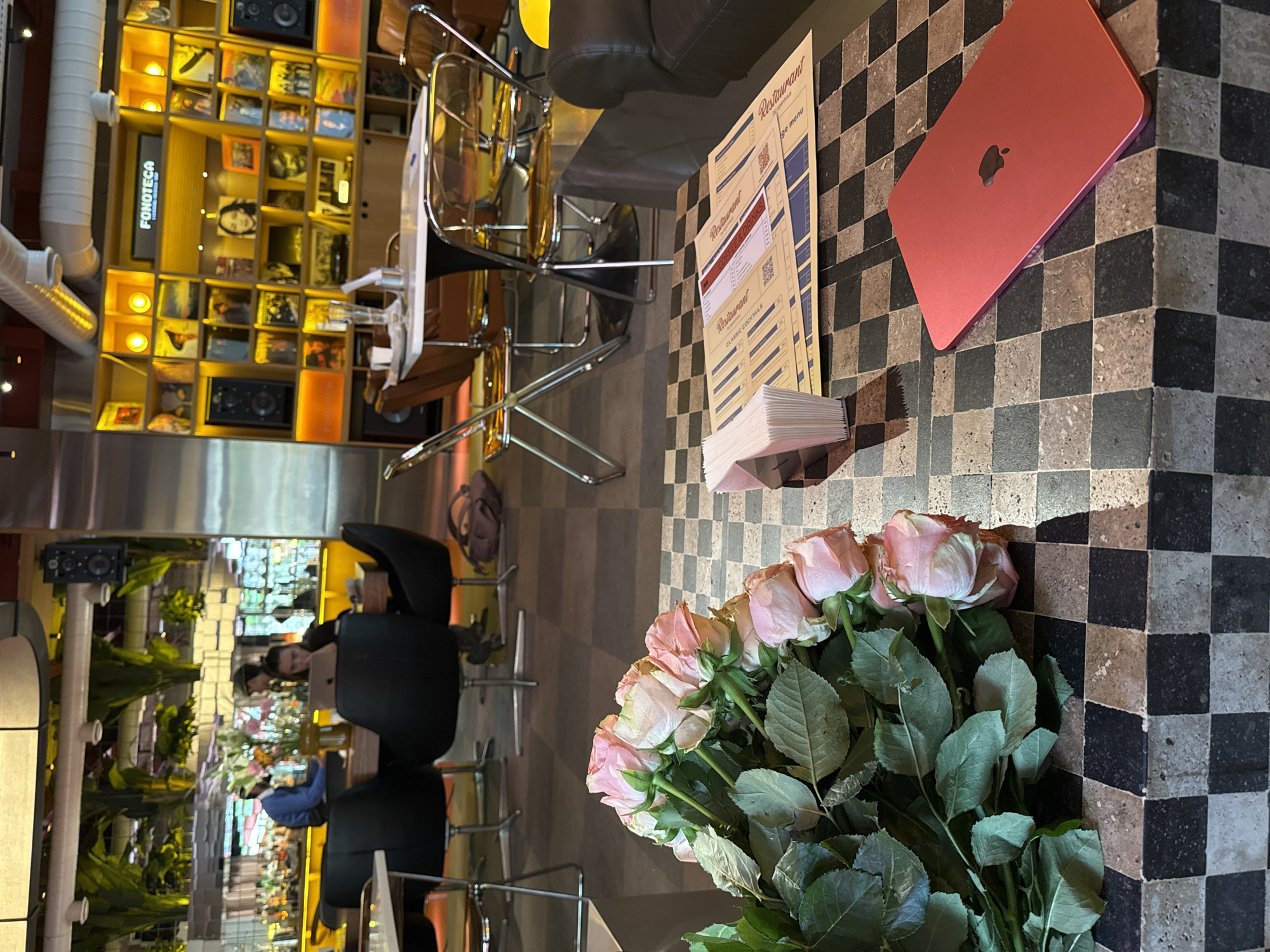 The image shows a table in a cafe or restaurant, likely in Yerevan, Armenia, as "FONOTECA" is visible on a shelf unit in the background. The table has a checkerboard pattern. A pink laptop, a stack of restaurant menus, and a large bouquet of pink roses are on the table. In the background, chairs and other tables are visible, along with people sitting and presumably eating or drinking. A reflective surface shows the restaurant's interior, including hanging plants and more seating. The lighting suggests it's either daytime or the cafe is well-lit with artificial lights.