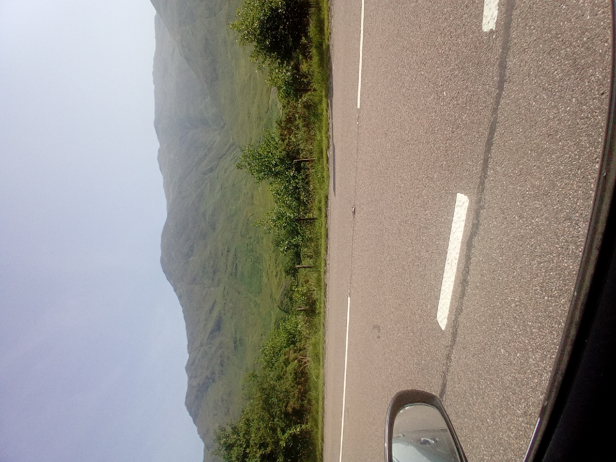 The image shows a view from a car window, looking out at a mountain landscape. The foreground is dominated by an asphalt road with a white line marking the lane. The road appears to be a two-lane highway. To the left, a portion of the car's side mirror and window frame is visible. Beyond the road, there is a strip of green vegetation, including trees and bushes, with a fence line running through it. In the background, a large, green, grass-covered mountain range rises into a pale blue, hazy sky. The overall impression is of a scenic drive through a rural, mountainous area. The location is specified as Kyle, United Kingdom, suggesting a setting in the Scottish Highlands.