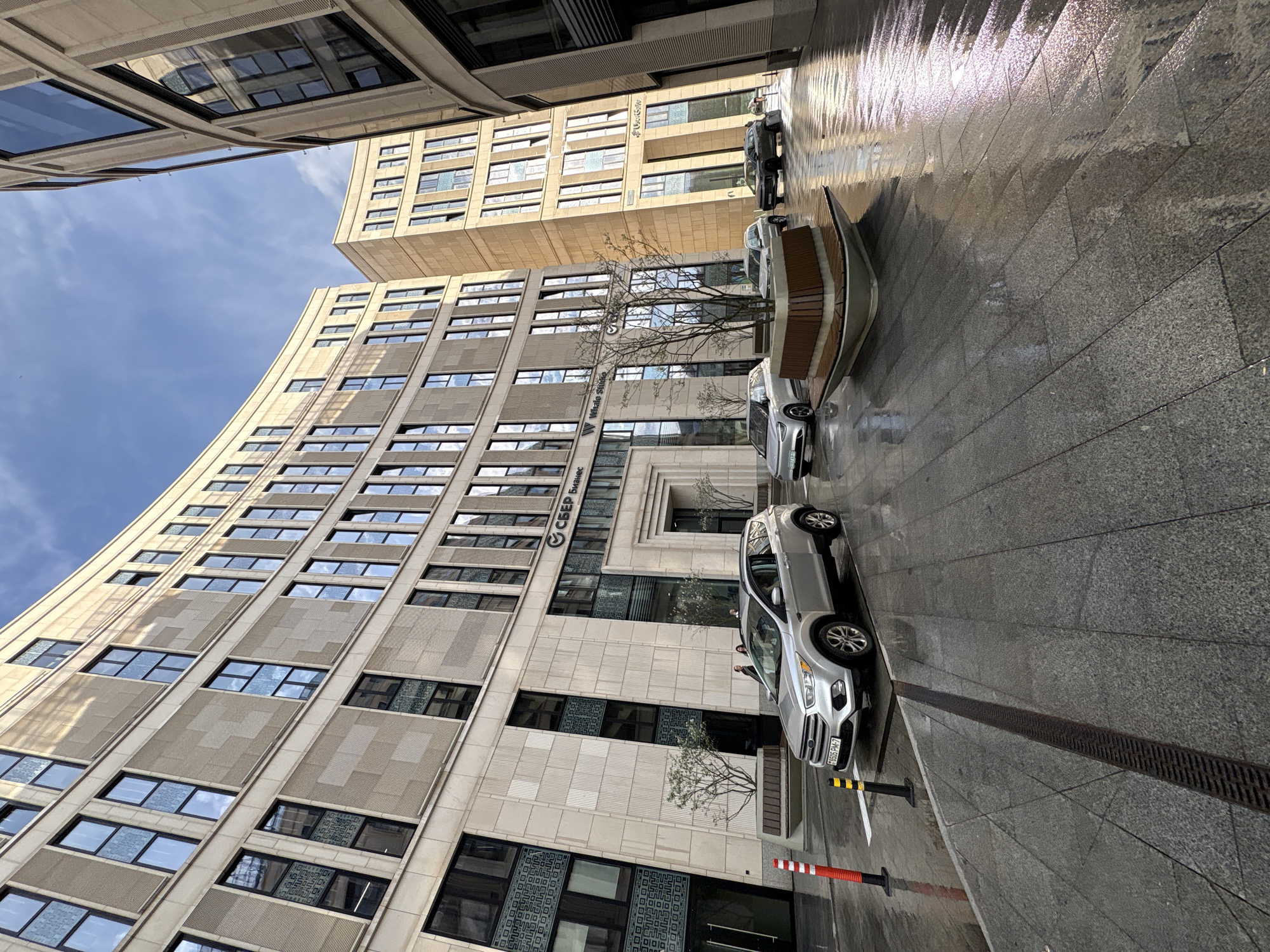 The image captures a street scene in Minsk, Belarus. A tall, modern building with numerous windows occupies a significant portion of the frame, its curved facade facing the viewer. Parked cars line the side of the street adjacent to the building, a silver Ford SUV prominently featured. The street surface appears wet and reflective, indicating recent rain. A pedestrian is slightly visible through the glass of the SUV. Street furniture, including a unique bench with a wooden top, is present. The sky is visible above the buildings, appearing partly cloudy. A sign that reads "SBER Business" in Cyrillic is visible on the building. In the foreground, red and white striped bollards mark the edge of the parking area. A metal drainage grate runs along the edge of the pavement.