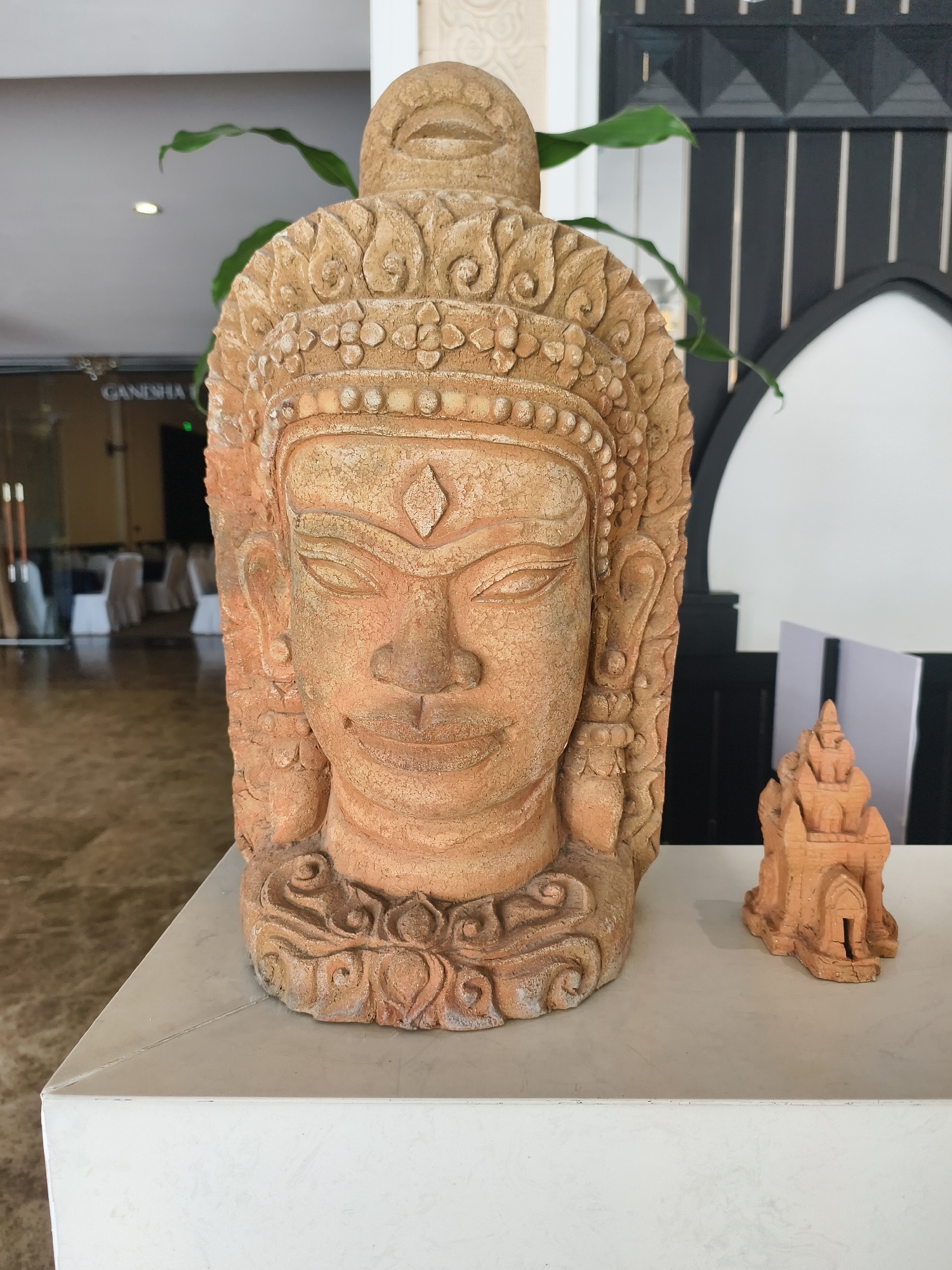 In Bac Nha Trang Ward, Vietnam, a close-up shot reveals a large, ornate terracotta head sculpture with intricate detailing of facial features, headwear, and neck adornments. To the right, a smaller terracotta replica of a building or temple sits beside the head. The sculptures are placed on a white marble-like surface. Background details suggest an interior setting with elements of decor and a partially visible room with tables and chairs.
