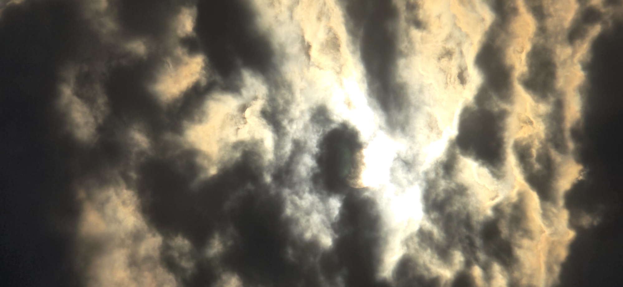 This is a vertical, close-up, low-angle photograph of a cloudy sky. The clouds are dark and ominous in the foreground and at the top, with brighter, golden-yellow clouds interspersed throughout the midground. The sun is visible, peeking through a gap in the clouds, creating a bright, hazy effect. The clouds are varied in texture and density, with some appearing wispy and others more solid. The overall impression is one of dramatic and turbulent weather. The location is in the city of Русская Деревня-2 Кп, Russia.