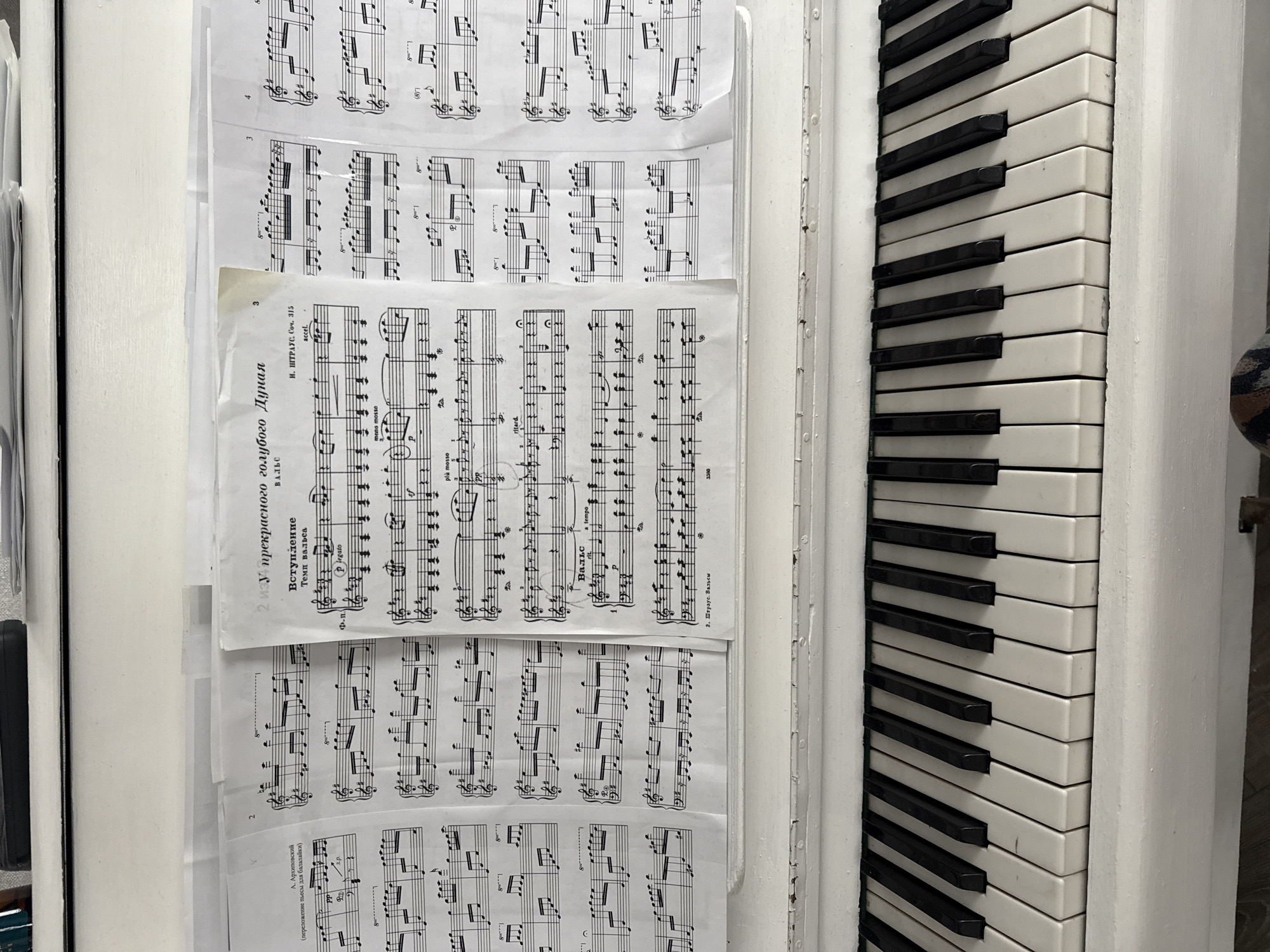 The image shows a white piano with sheet music spread across the keys. The sheet music is in Russian and appears to be classical music, with titles such as "Вступление" (Introduction) and "Вальс" (Waltz). The piano is positioned in what seems to be an indoor setting, possibly a home or a music studio, though no specific location details are visible. The piano is old and shows signs of wear and tear, with chipped paint on its surface. The lighting suggests it is daytime.