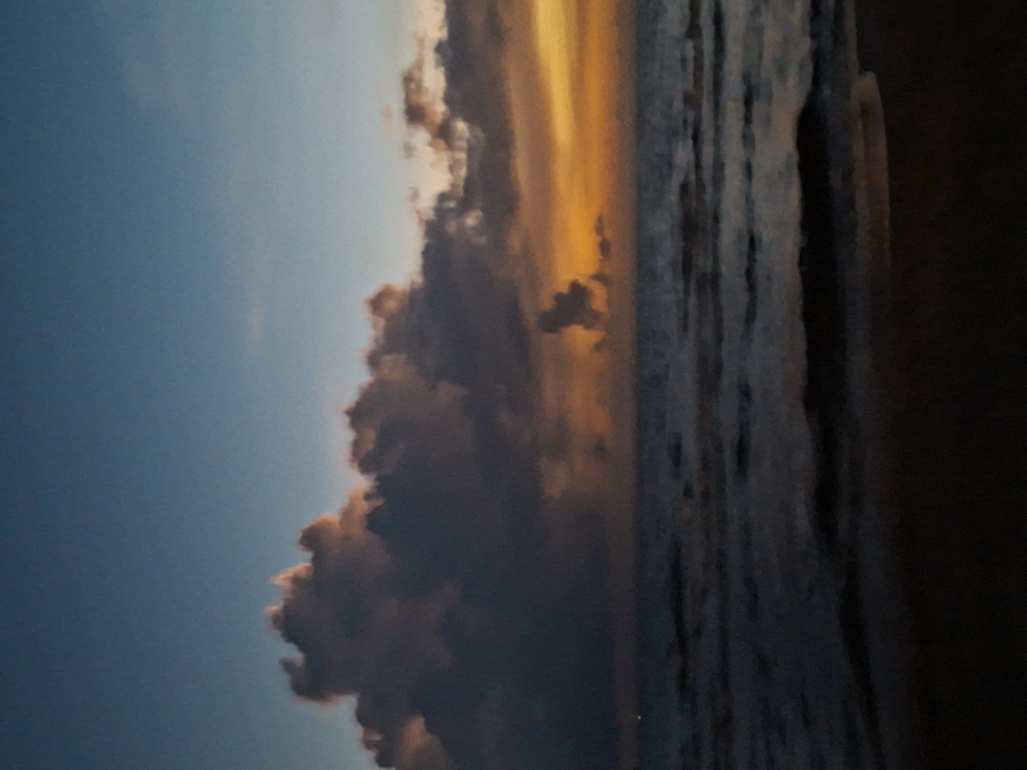 The image depicts a serene seascape at dusk. A dark, textured shoreline occupies the bottom portion of the frame, transitioning into the ocean. The water is dark and somewhat choppy, with visible waves. Above the water, dramatic clouds dominate the sky. These clouds are a mixture of dark grays and purples, with vibrant streaks of orange and yellow illuminating them from behind, suggesting the setting sun. The upper portion of the sky is a deep, clear blue. There are no people, structures, or distinct man-made objects visible in the scene. The overall impression is one of natural beauty and tranquility during twilight.