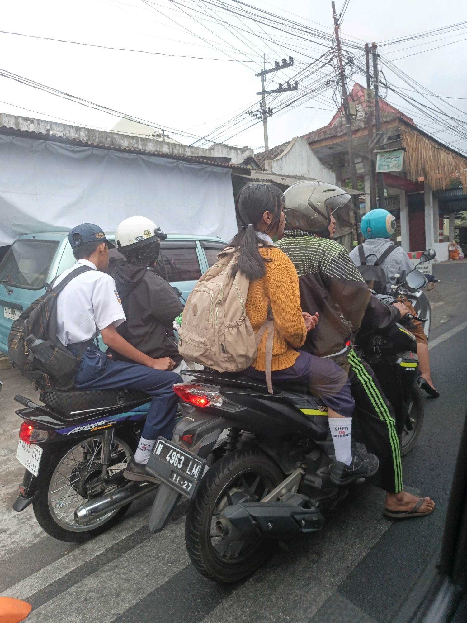 The image captures a street scene in Batu, Indonesia, with several motorcycles and people. The foreground features the rear of two motorcycles, one with a license plate reading "N 4963 LM 11-27." A person wearing white socks with "SNPN 03 KOTA BATU" printed on them is visible. On the left motorcycle, a student in a white shirt and blue pants, wearing a blue cap and carrying a backpack, is seated. Beside them is another person wearing a black jacket and a white helmet.
In the mid-ground, a motorcycle carries three people. The person in front is wearing a checkered shirt and a grey helmet. The person in the middle is wearing a yellow knitted sweater and a beige backpack. The person at the back is wearing a grey shirt and a blue helmet. These individuals appear to be students given their attire.
The background shows buildings with corrugated iron roofs and awnings, typical of a local Indonesian urban setting. A light blue car is parked to the left. Overhead, a complex network of electrical wires hangs from poles, indicating an urban environment with significant infrastructure. The sky is overcast, suggesting a cloudy day. The motorcycles are stopped on a road, possibly at an intersection or traffic light, as indicated by the crosswalk lines on the road surface.